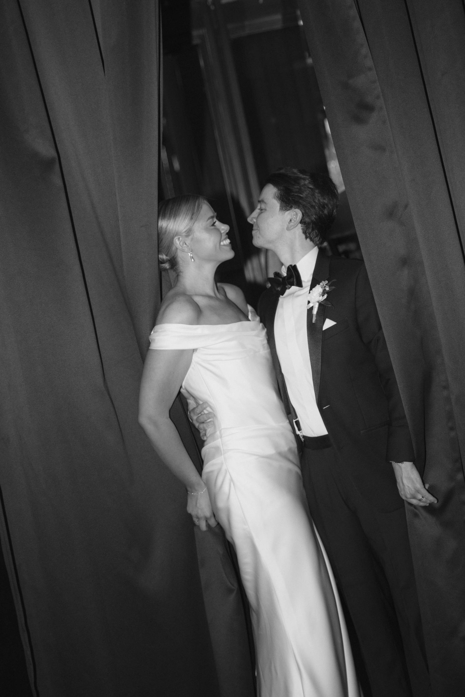 Black and white photo of a bride and groom sharing a romantic moment between dark curtains at their wedding.