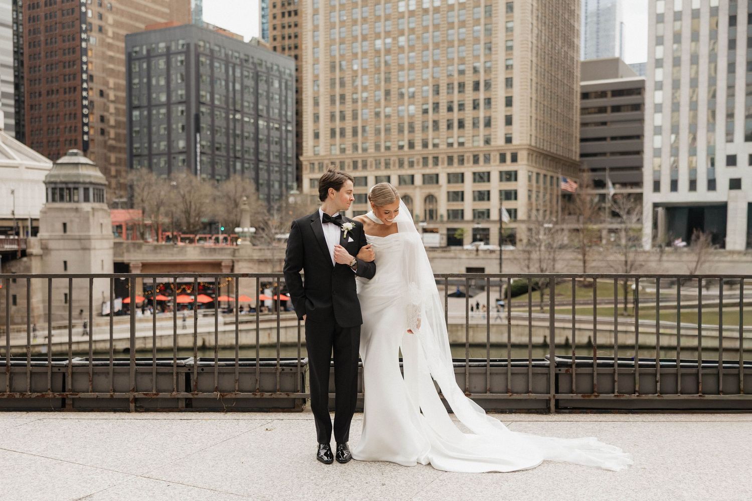 Bride and groom pose on Chicago Riverwalk with city skyline backdrop, bride in elegant white gown with long veil.
