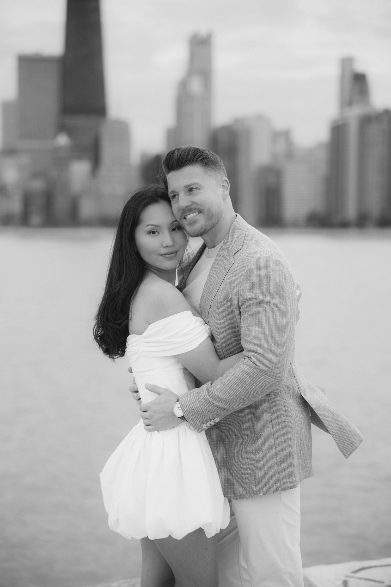 Black and white photo of a couple embracing by the Chicago skyline waterfront, woman in white dress.