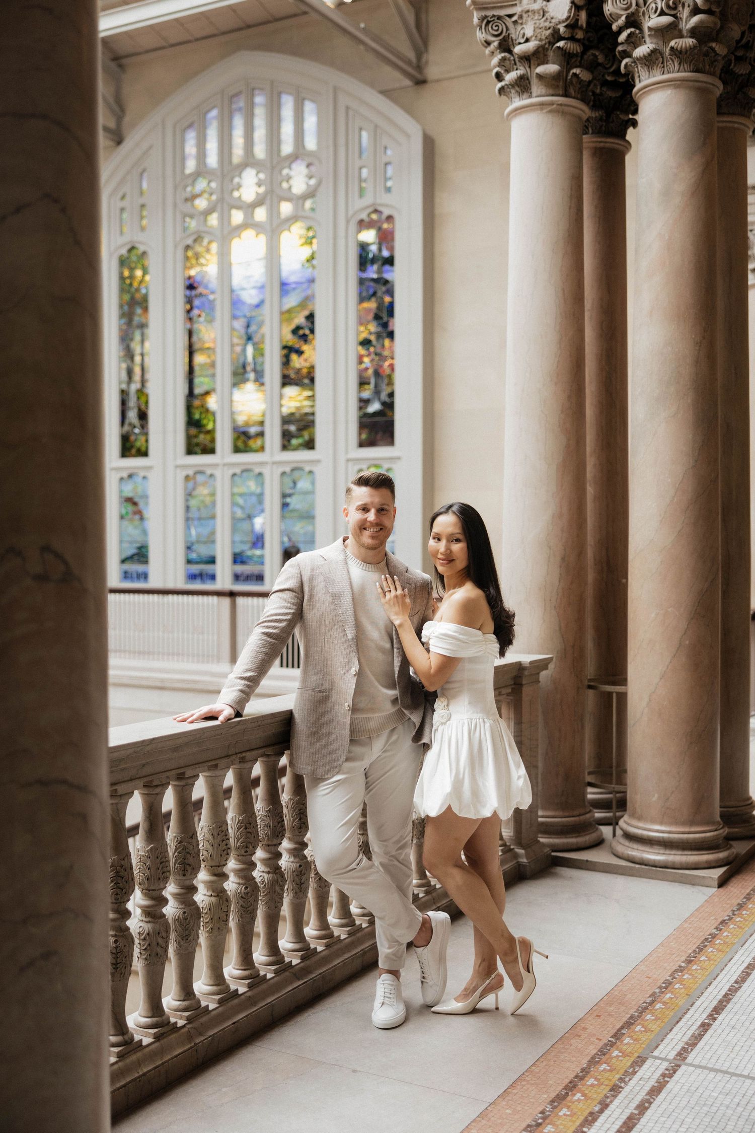 Couple posing in elegant hall with stained glass window, woman in white dress showing engagement ring, man in beige suit.