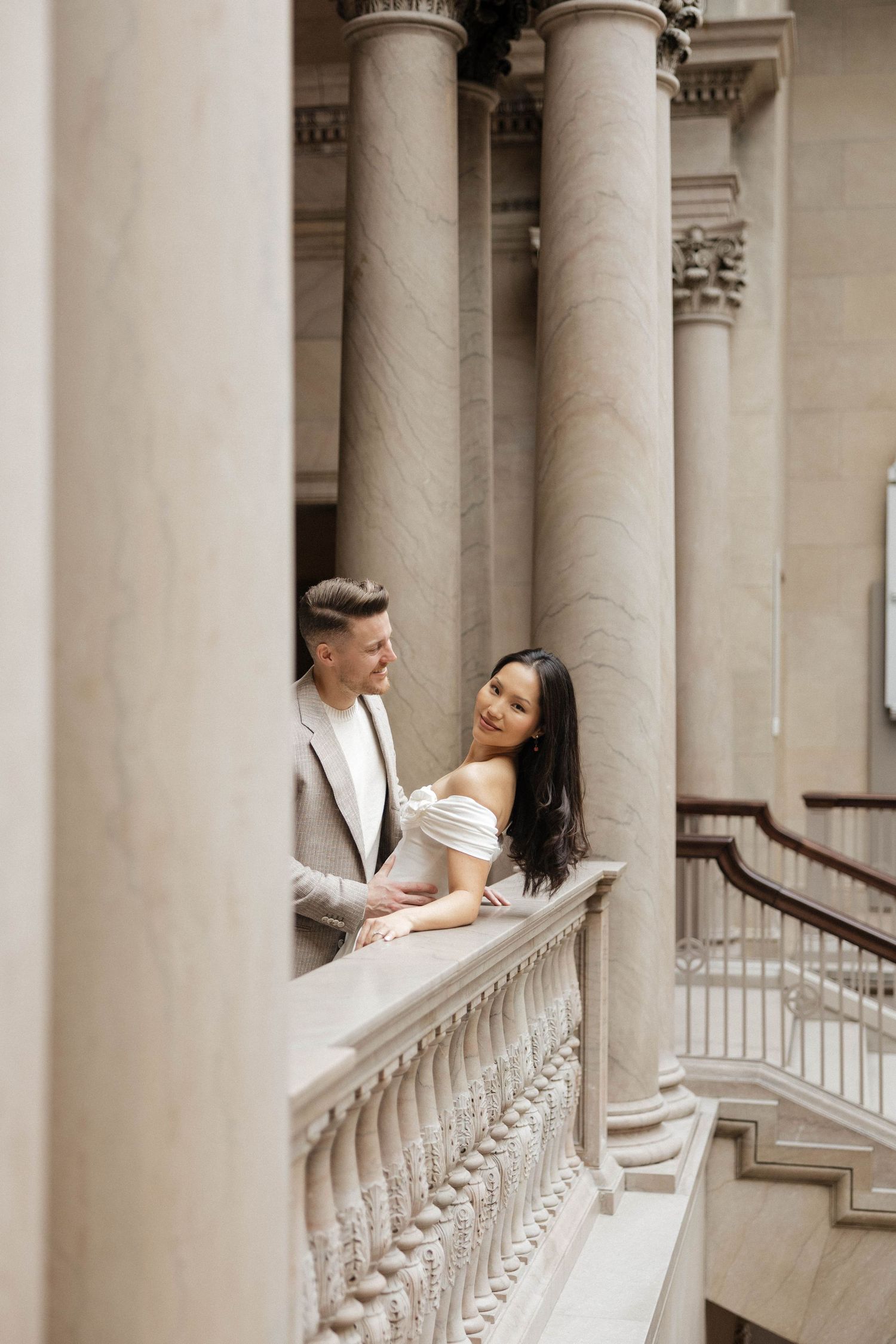 Elegant couple posing on a classical stone balcony with tall marble columns in a grand architectural setting.