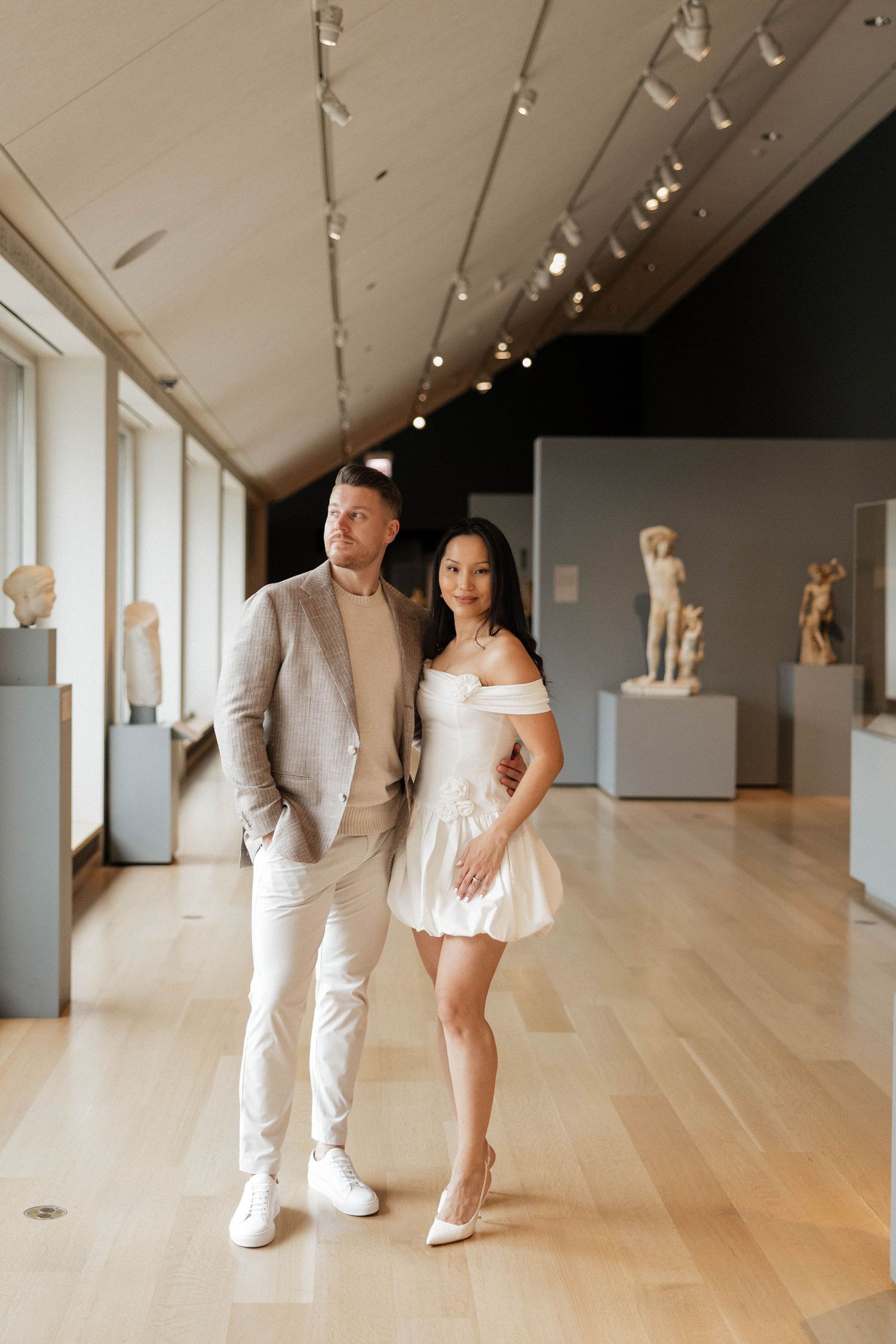 Elegant couple posing in art museum gallery with classical sculptures, man in beige suit, woman in white mini dress.