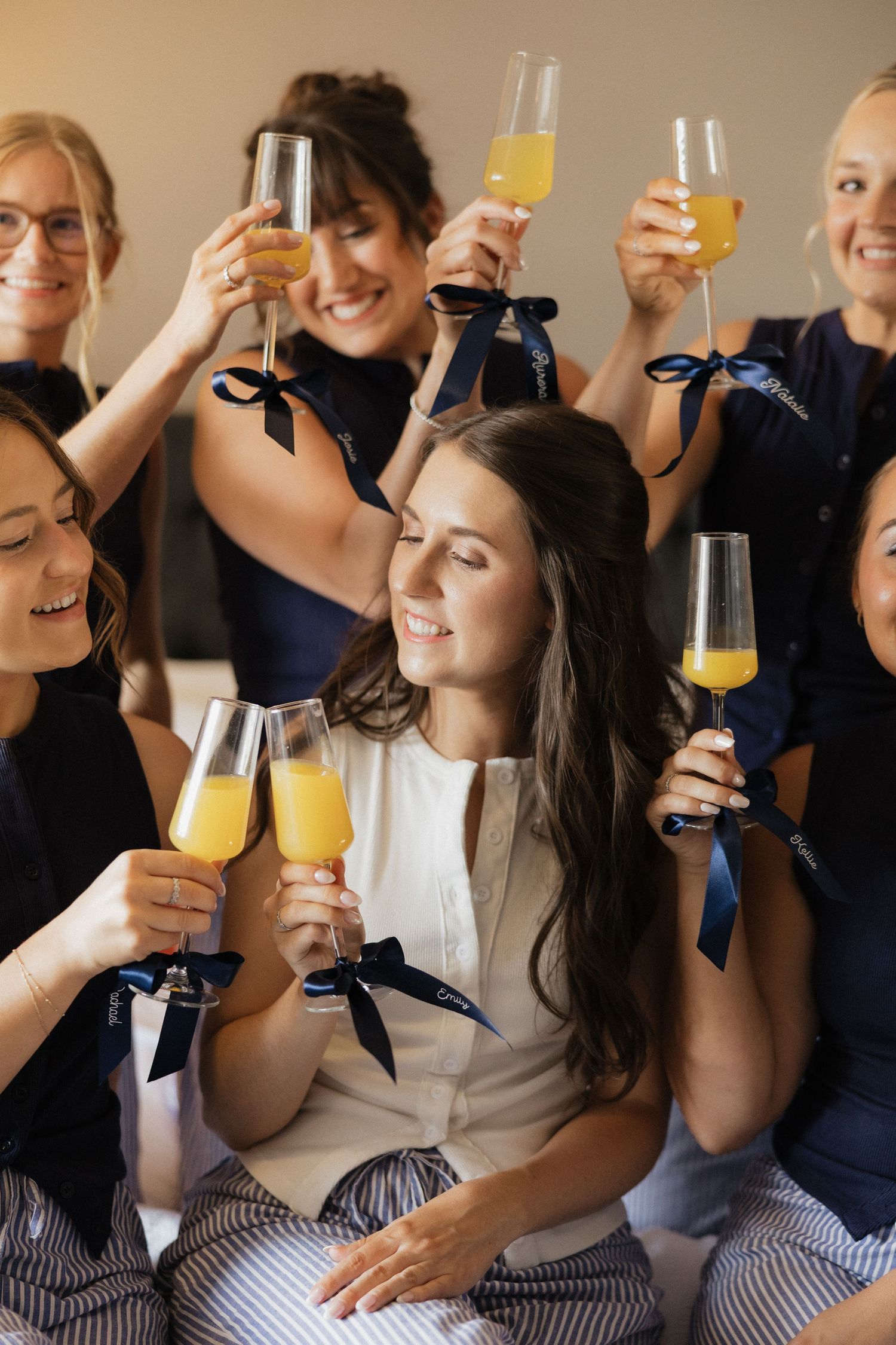 Bridesmaids in navy robes toasting with mimosas alongside bride in white during wedding morning preparations.