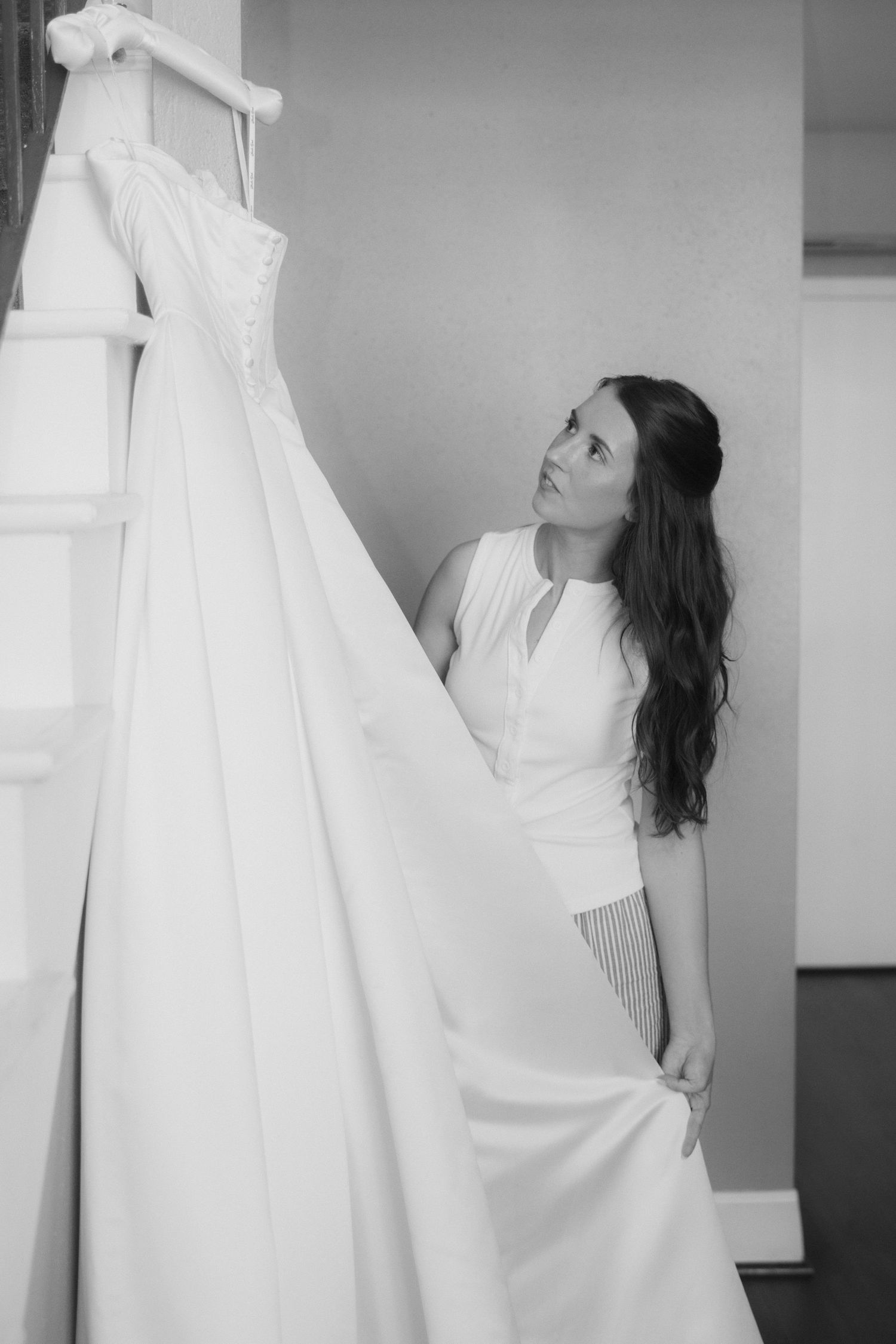 Bride in white dress admiring her wedding gown hanging on staircase in black and white photo.