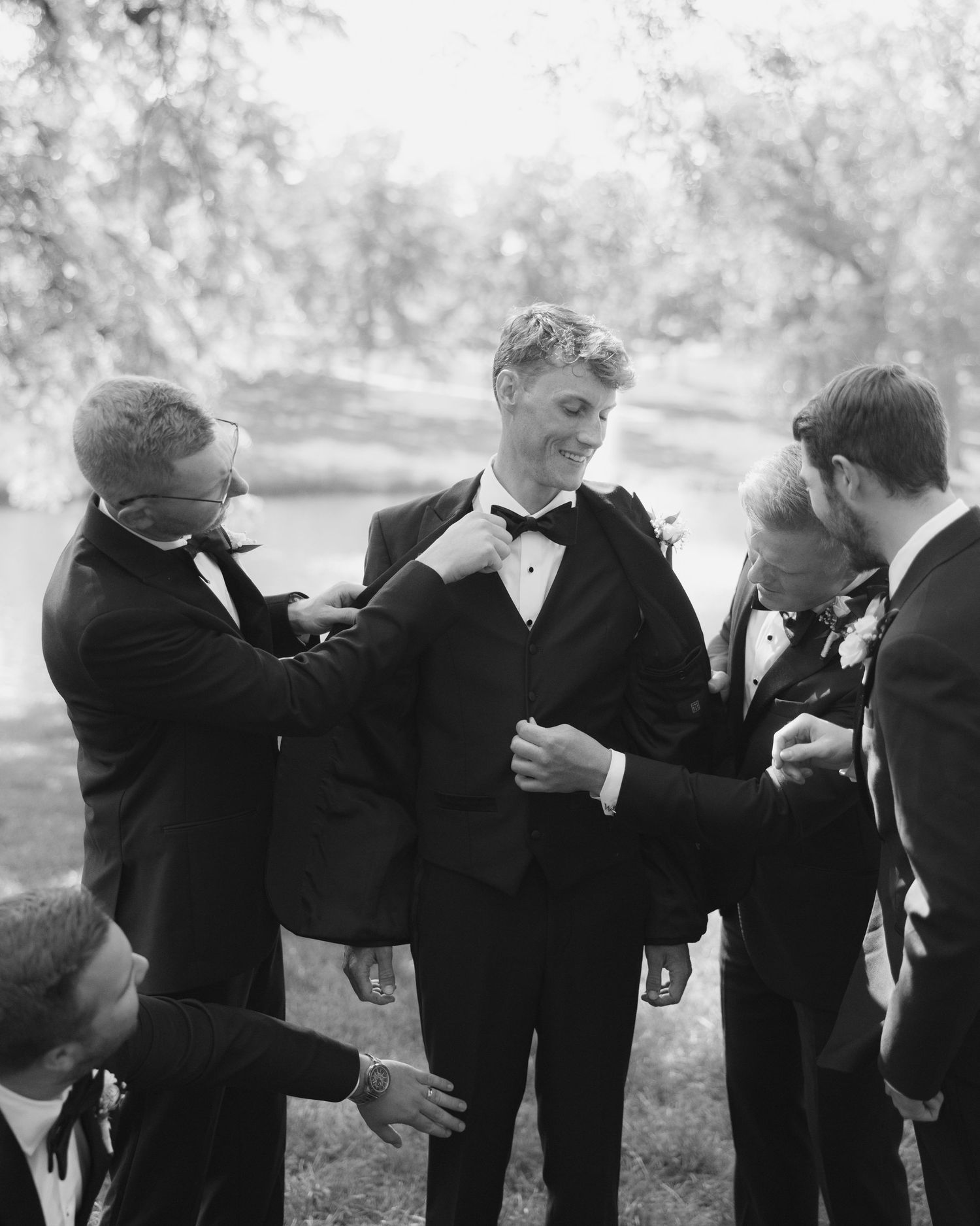 Groomsmen in black tuxedos help the groom prepare outdoors in a classic black and white wedding photo.