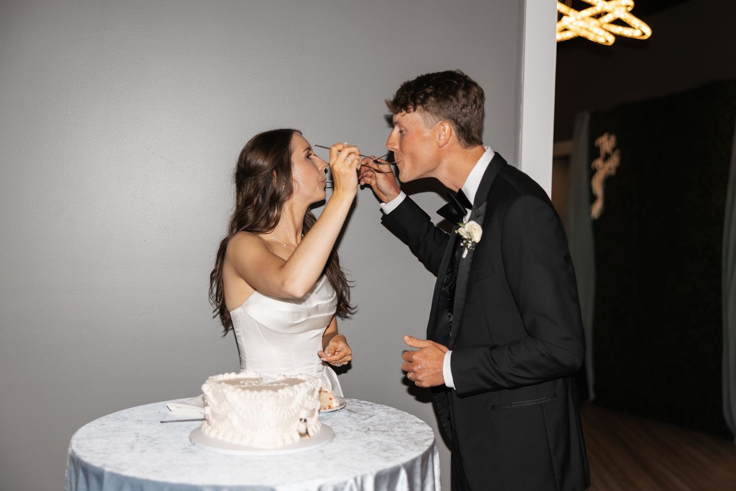 Bride and groom sharing wedding cake at reception, dressed in white gown and black tuxedo, smiling together.