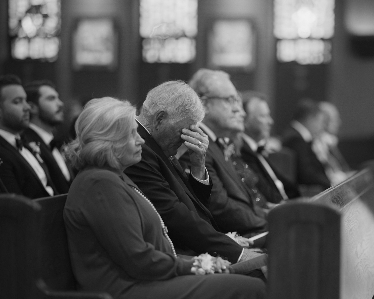 Black and white photo of congregation sitting in church pews during a solemn ceremony, elderly man bowing head in prayer.