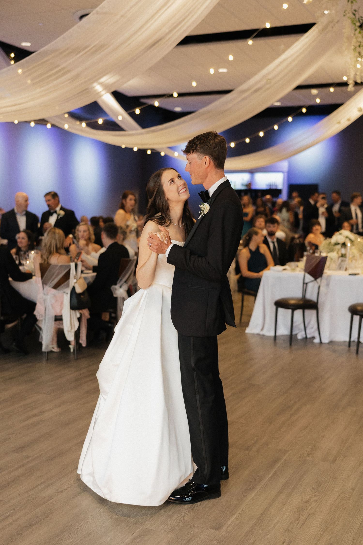 Bride and groom share their first dance at elegant wedding reception with string lights and draped ceiling decor.