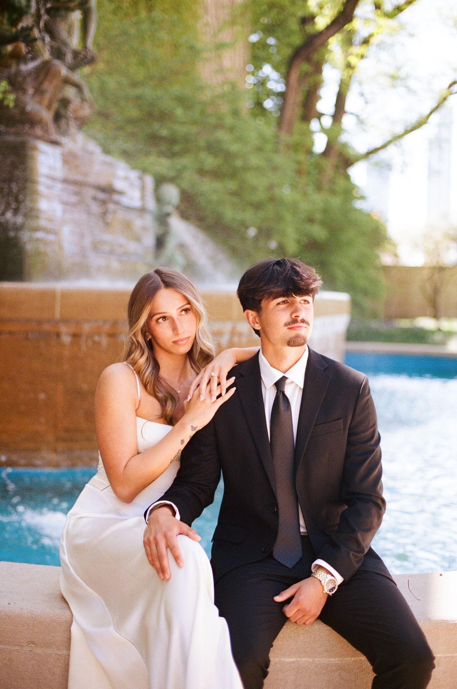 Couple in elegant wedding attire sitting by a fountain, woman in white dress resting hand on mans shoulder.