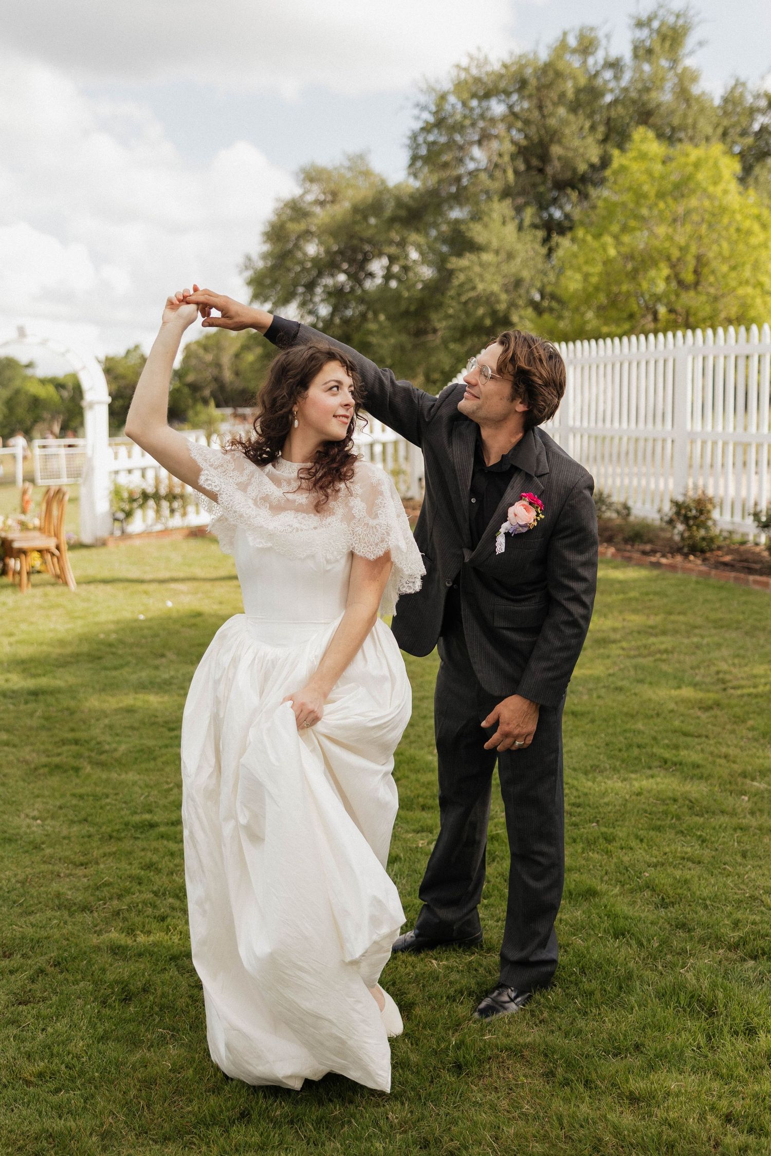 Bride in white lace gown twirls as groom in black suit spins her during outdoor wedding dance on green lawn.
