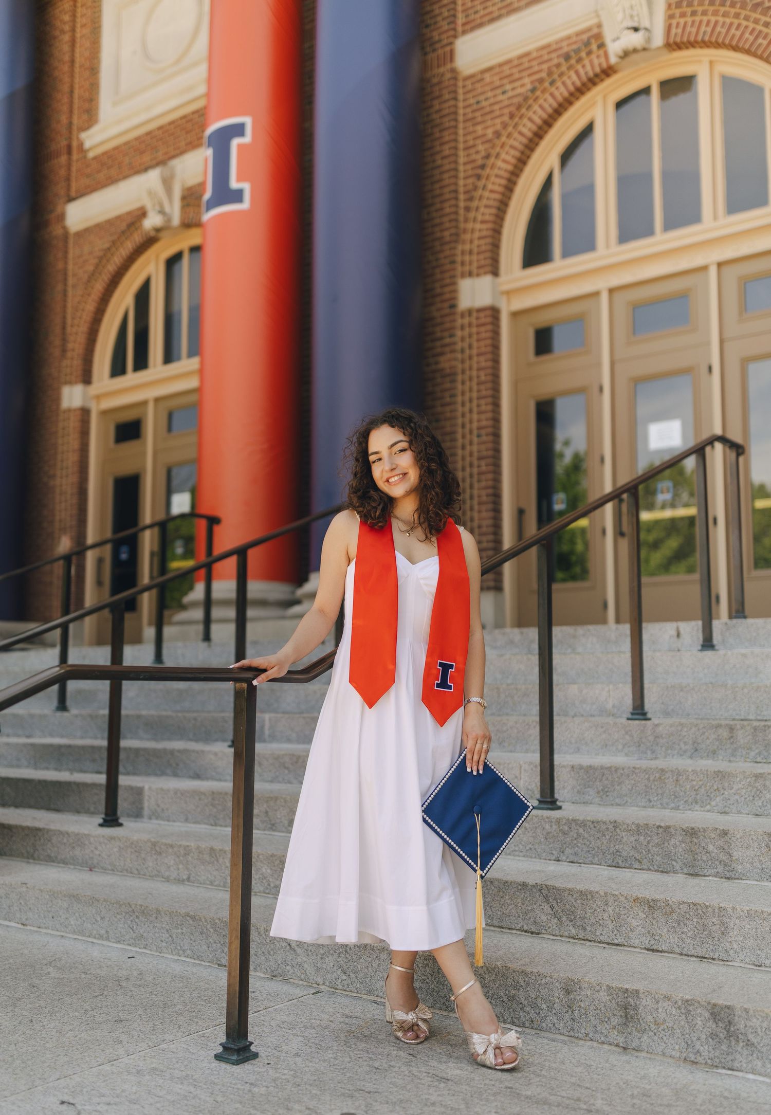 Graduate in white dress and orange stole poses on steps of brick university building with columns.
