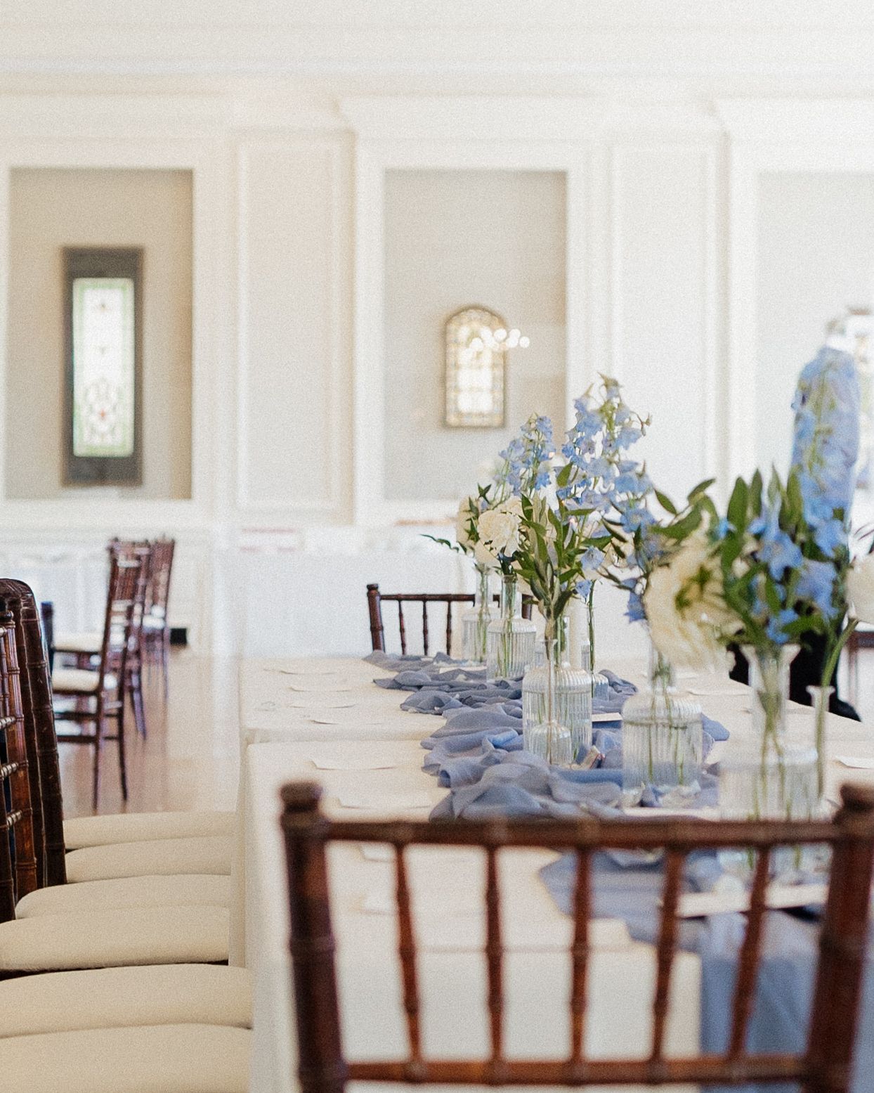 Elegant wedding reception setup with white tablecloths, dark wooden chairs, and delicate floral centerpieces in a bright room.