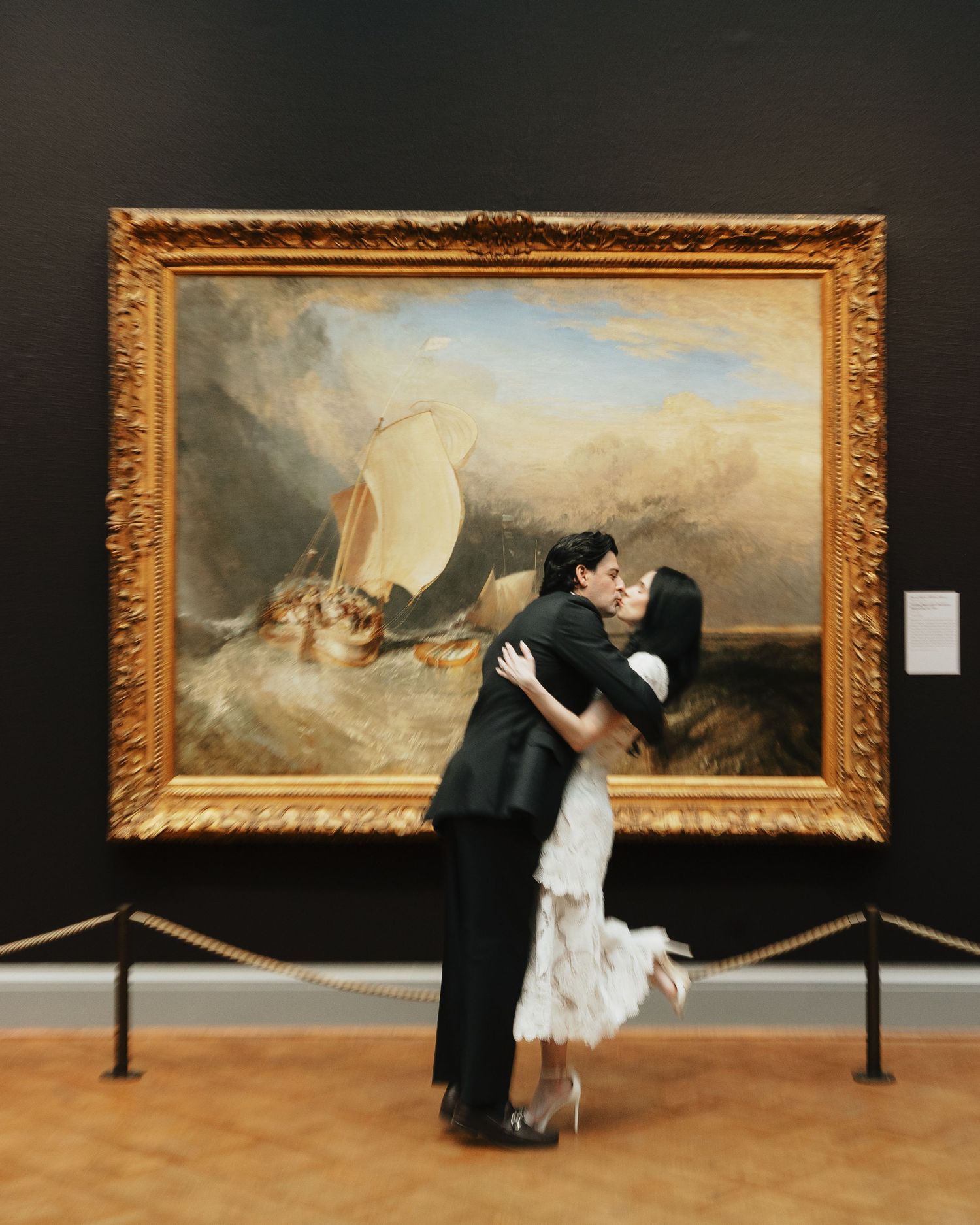 A couple in formal attire shares a kiss in an art gallery in front of a gilded-frame seascape painting of sailboats on stormy waves.