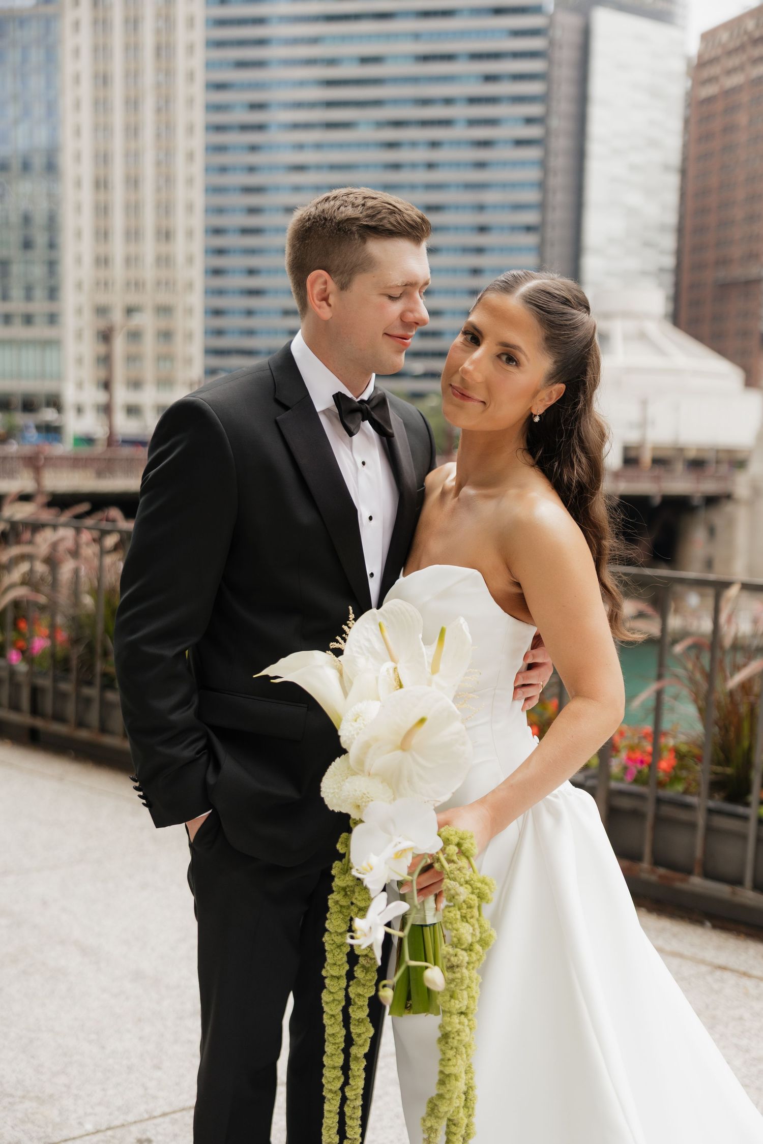 Bride in a strapless white gown holding a white calla lily and green amaranthus bouquet beside a smiling groom in a black tuxedo and bow tie