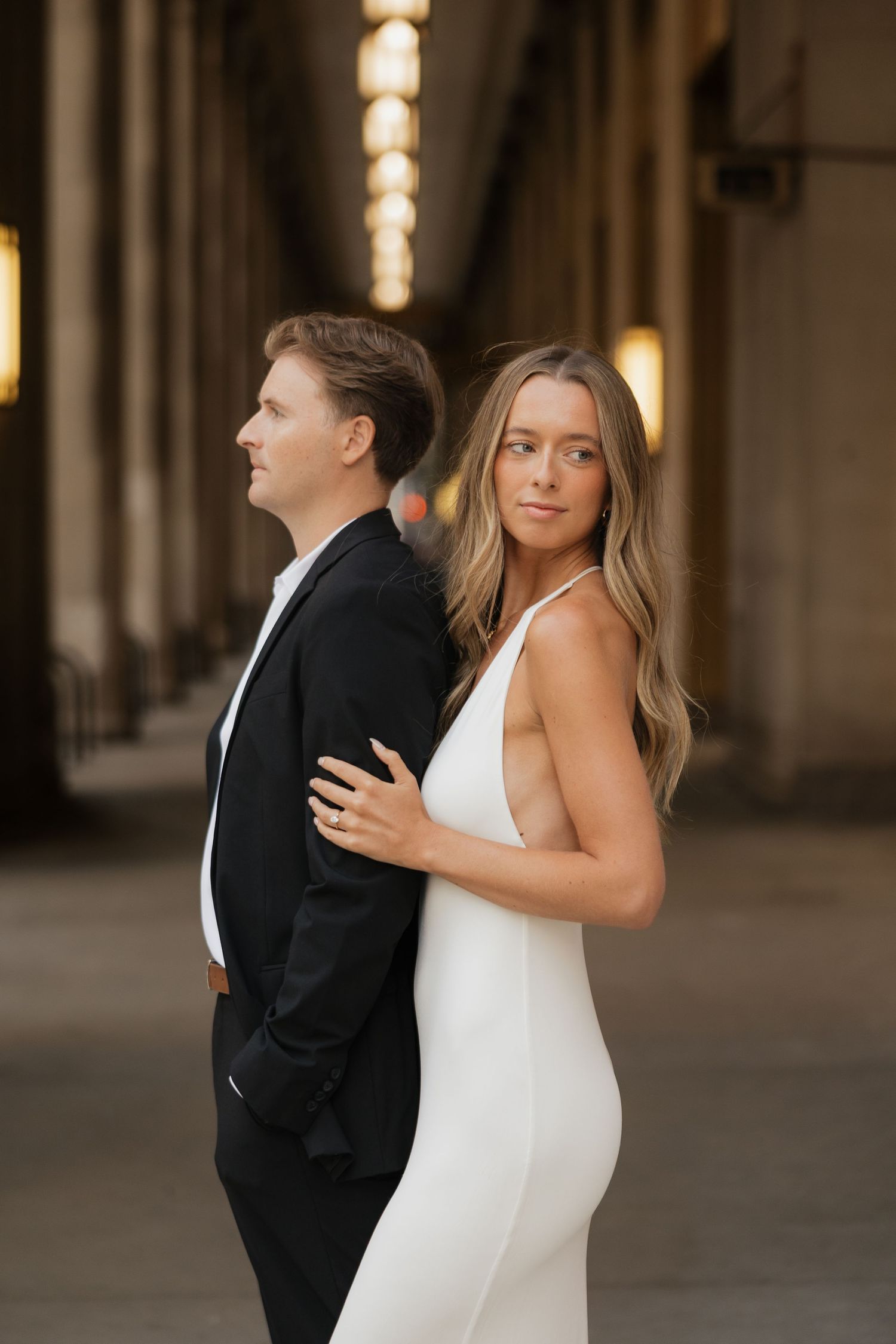 Couple standing back to back in a columned corridor at dusk, the man in a black suit and the woman in a white dress.