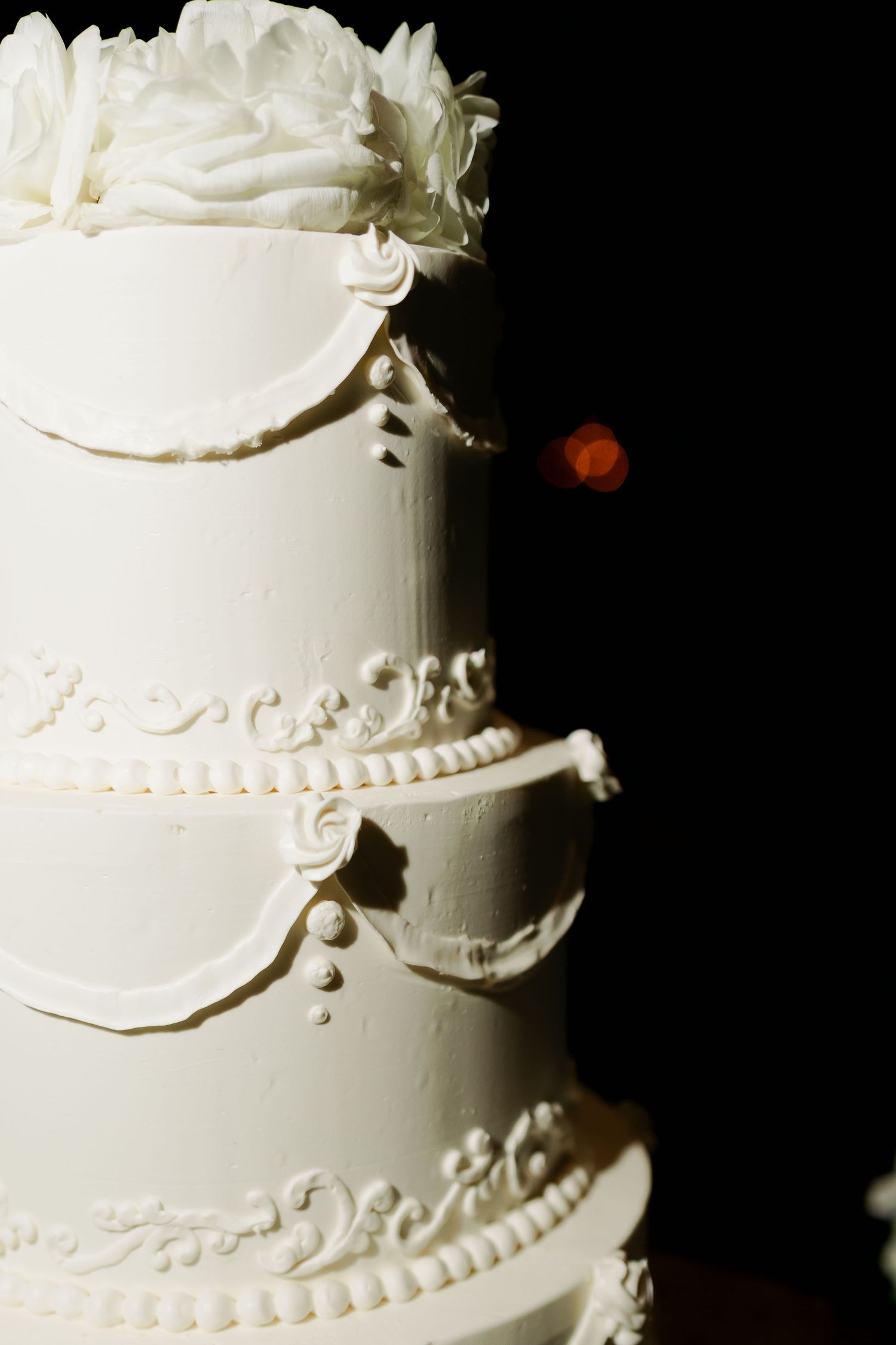 Elegant white tiered wedding cake with buttercream scrollwork, piped pearl borders, and a ruffled rose topper against a dark background.
