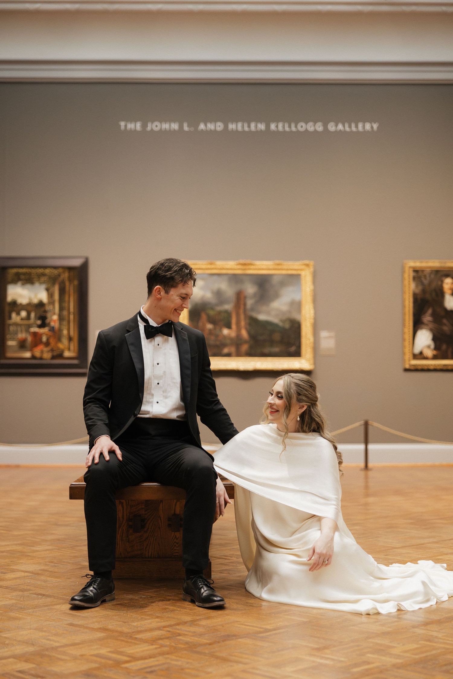 A couple in formal wedding attire sits together on a wooden bench in an art gallery with paintings on display.