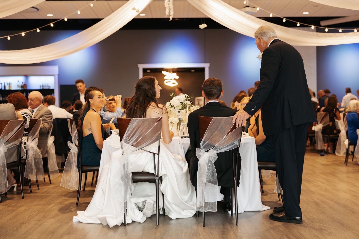 Wedding reception scene with bride and groom seated at head table while officiant speaks in elegant draped venue.