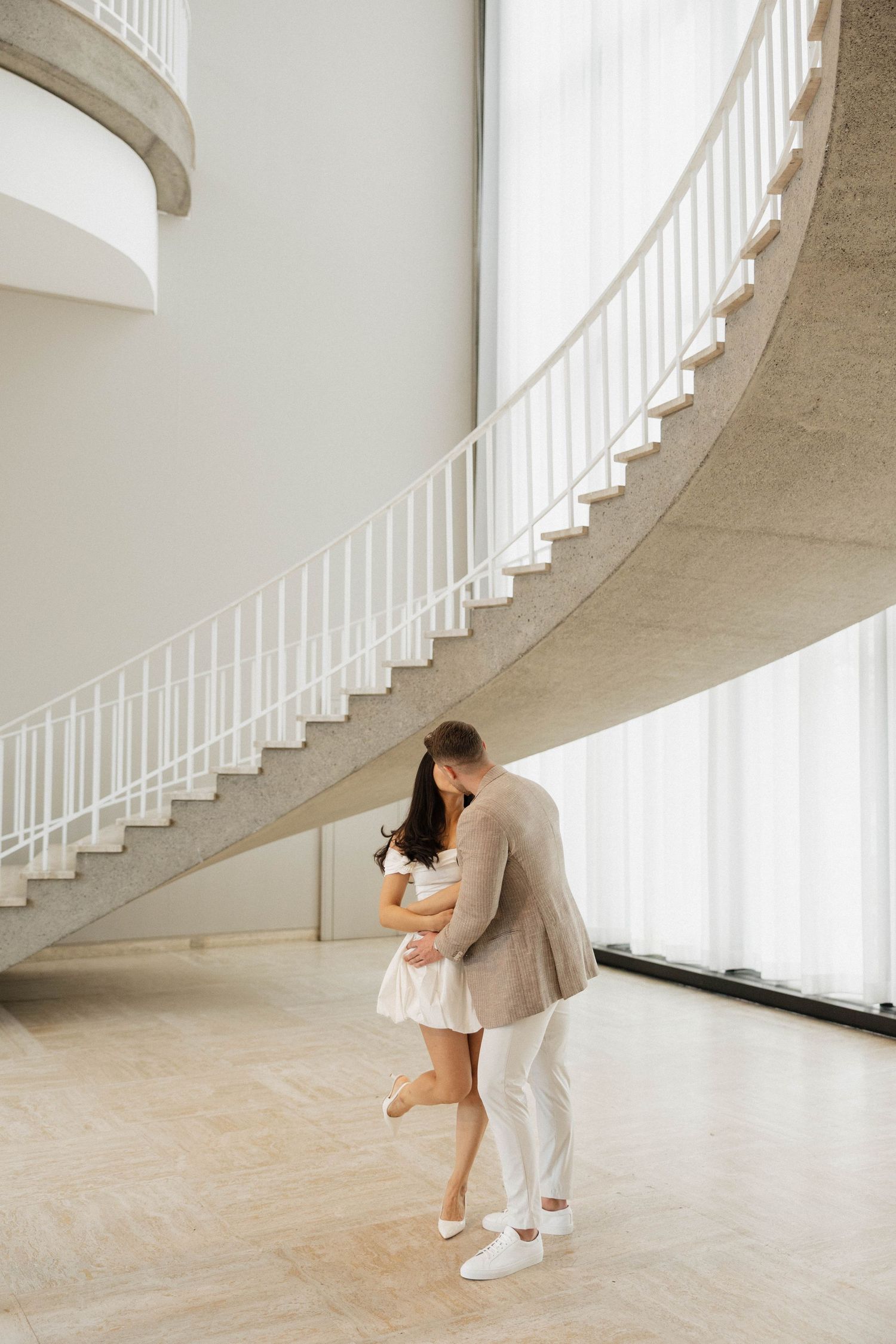 Couple embracing romantically beneath a curved spiral staircase in a bright modern building interior.