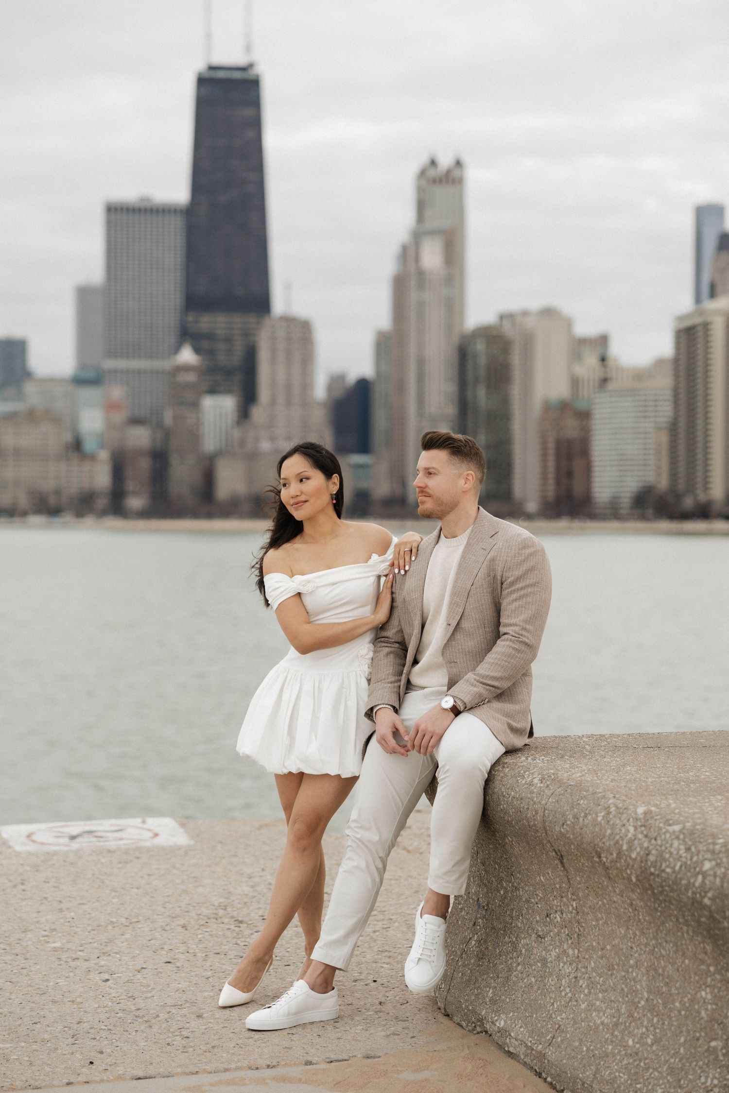 Couple posing by Lake Michigan with Chicago skyline backdrop, woman in white dress, man in beige suit.