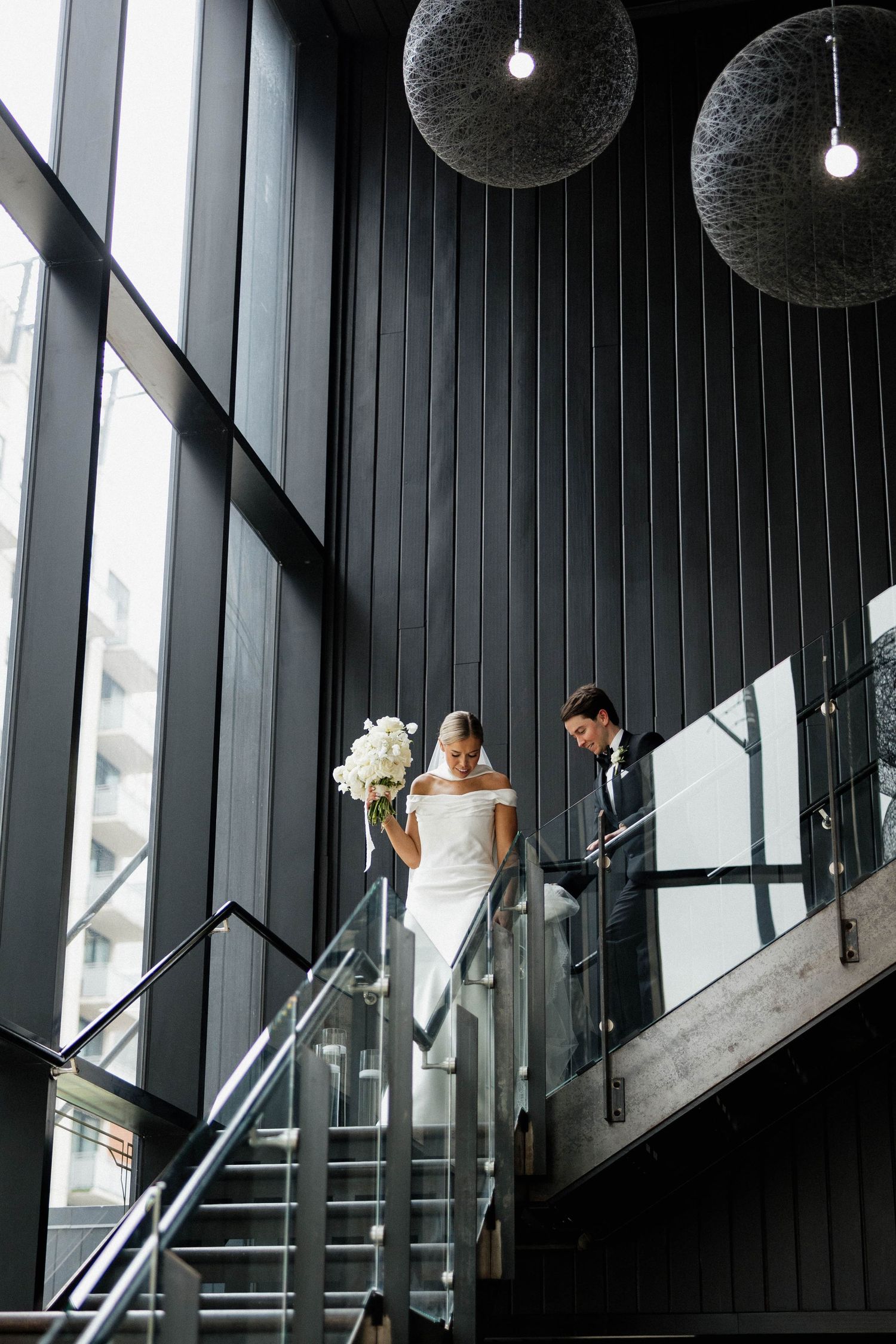 Bride and groom descend modern glass staircase, bride holding white bouquet, pendant lights above in elegant venue.