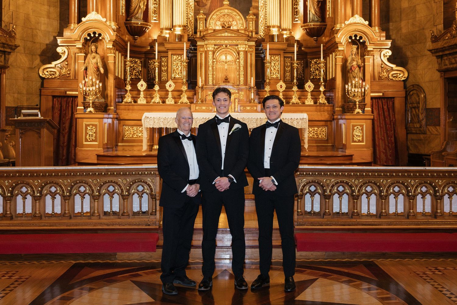 Groomsmen in formal attire stand together before decorative church altar with golden details.