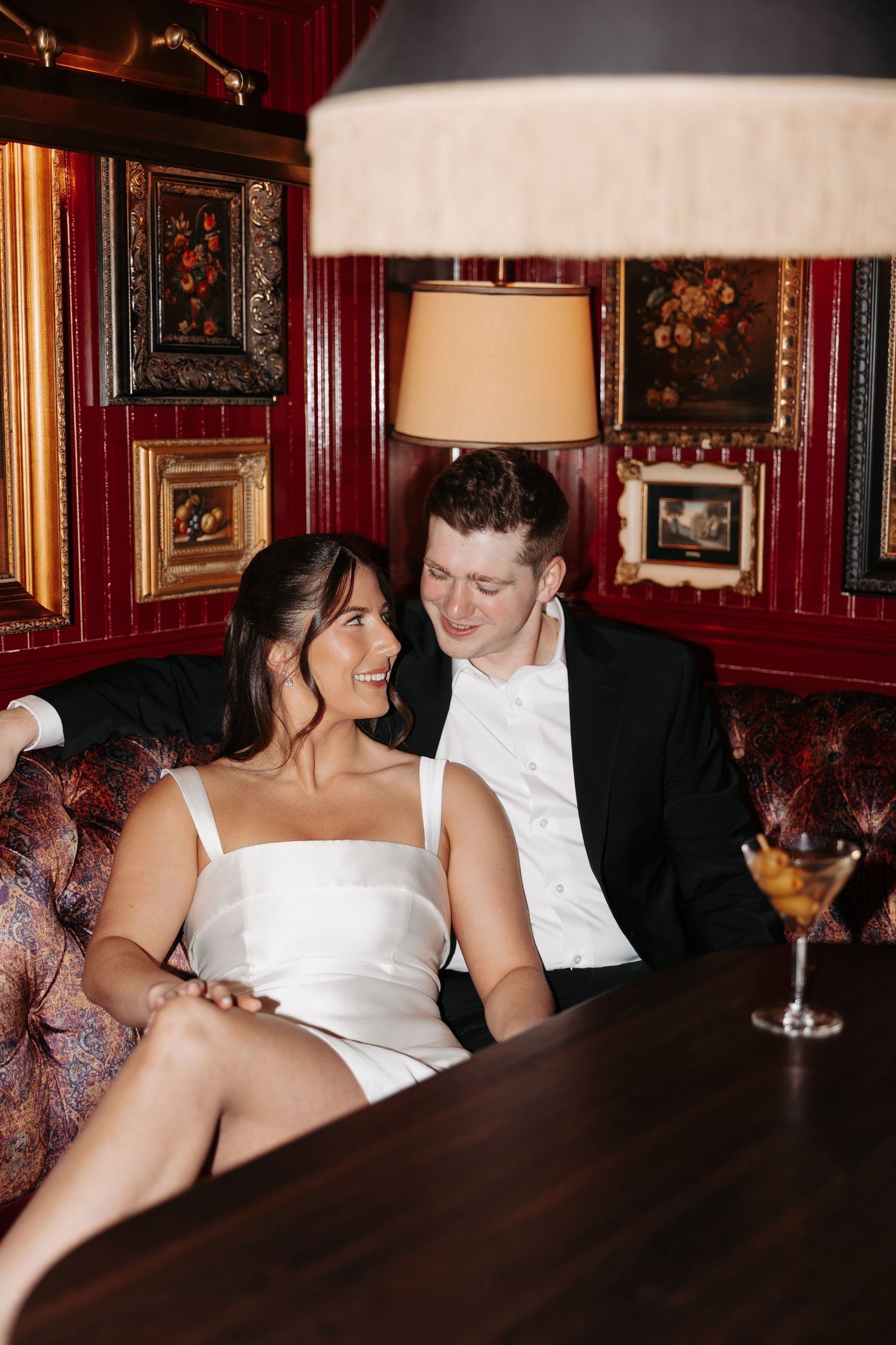 A couple laughs together while seated at a dark wood table in an elegant bar with red walls and framed artwork.