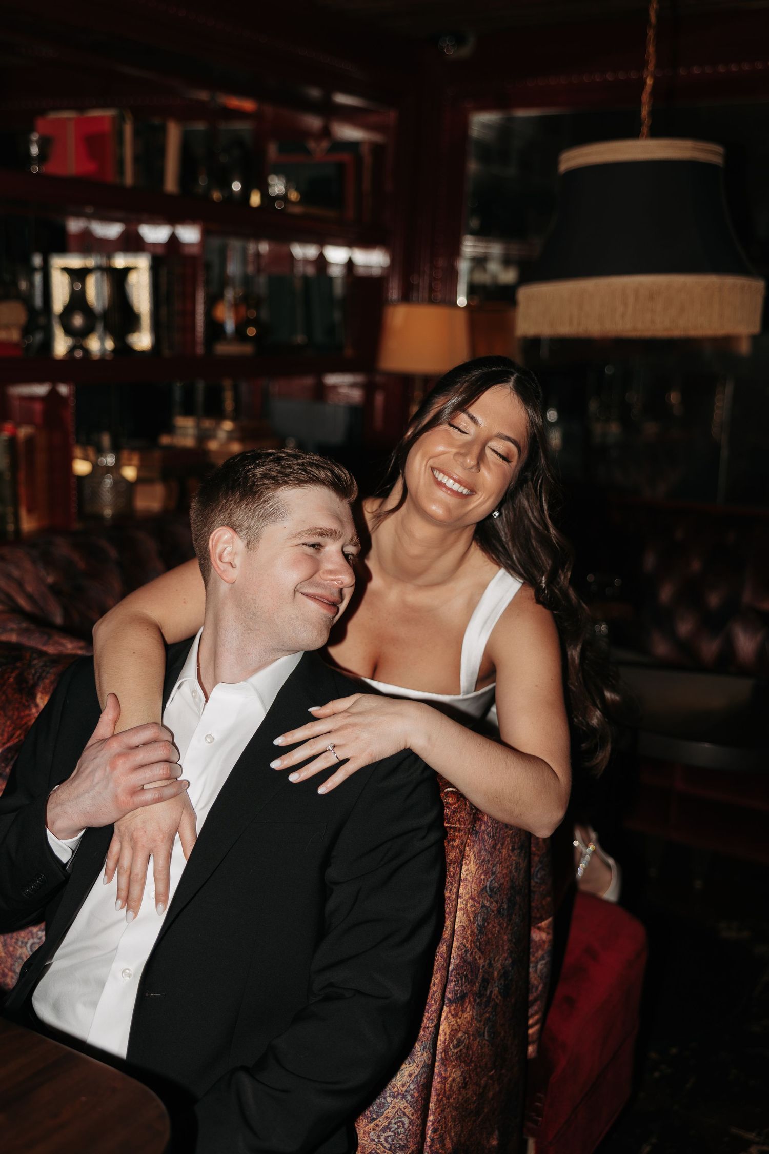 A couple shares intimate moments and laughter in formal attire at an indoor venue with dark ambient lighting.
