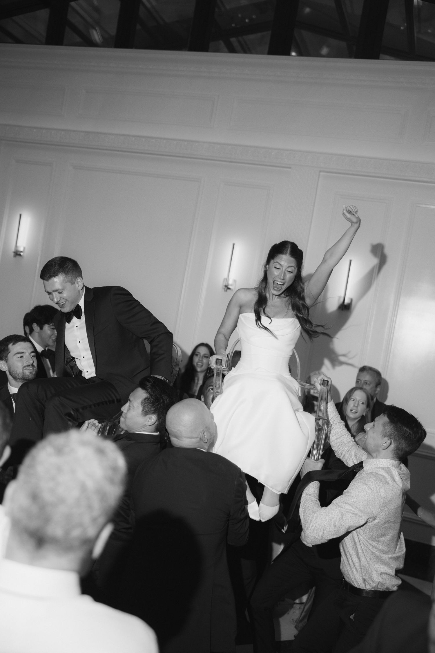 Bride and groom lifted by guests during joyful hora dance at elegant wedding reception in black and white photo.
