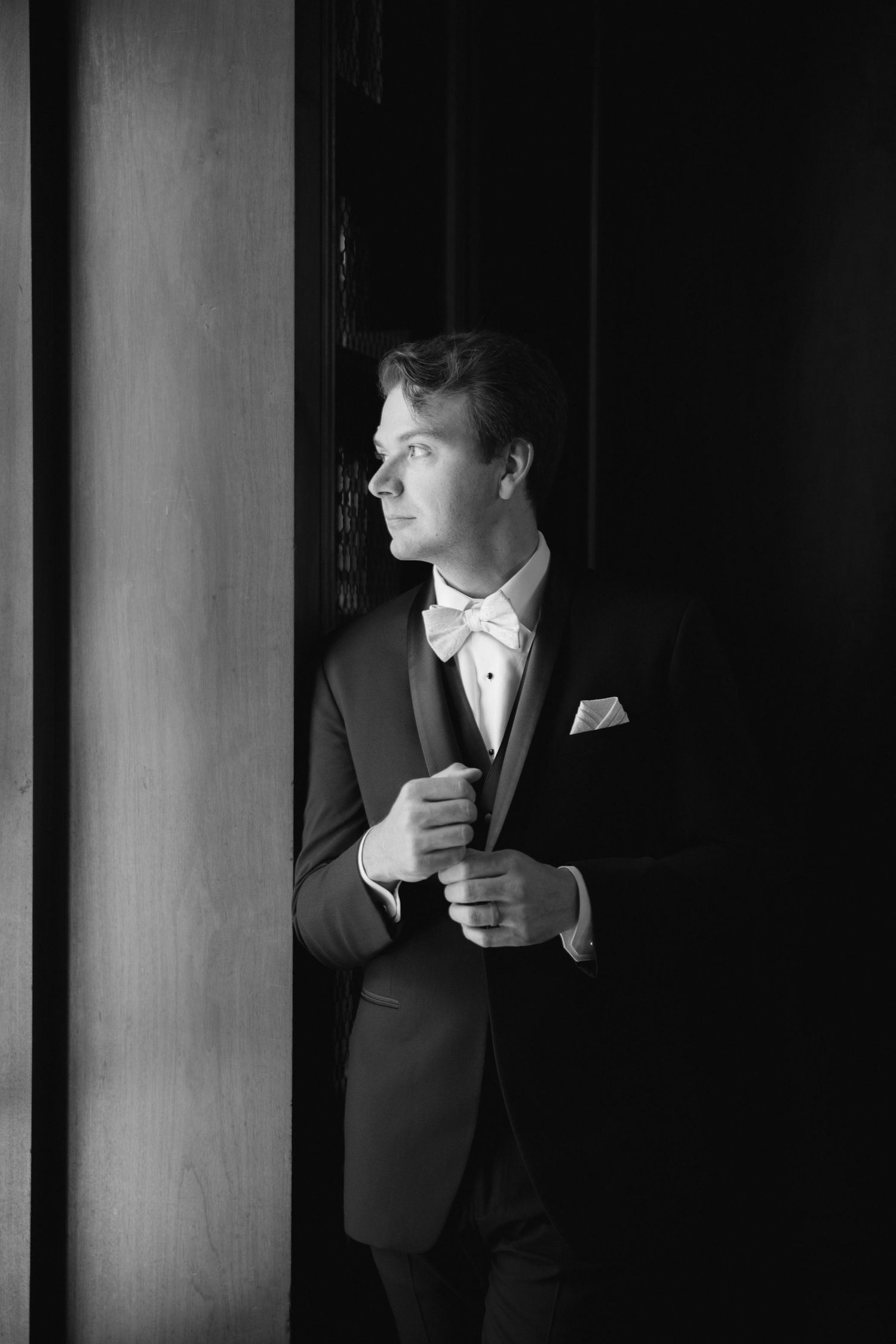 Black and white portrait of a dapper groom in tuxedo and bow tie standing by a dark doorway.
