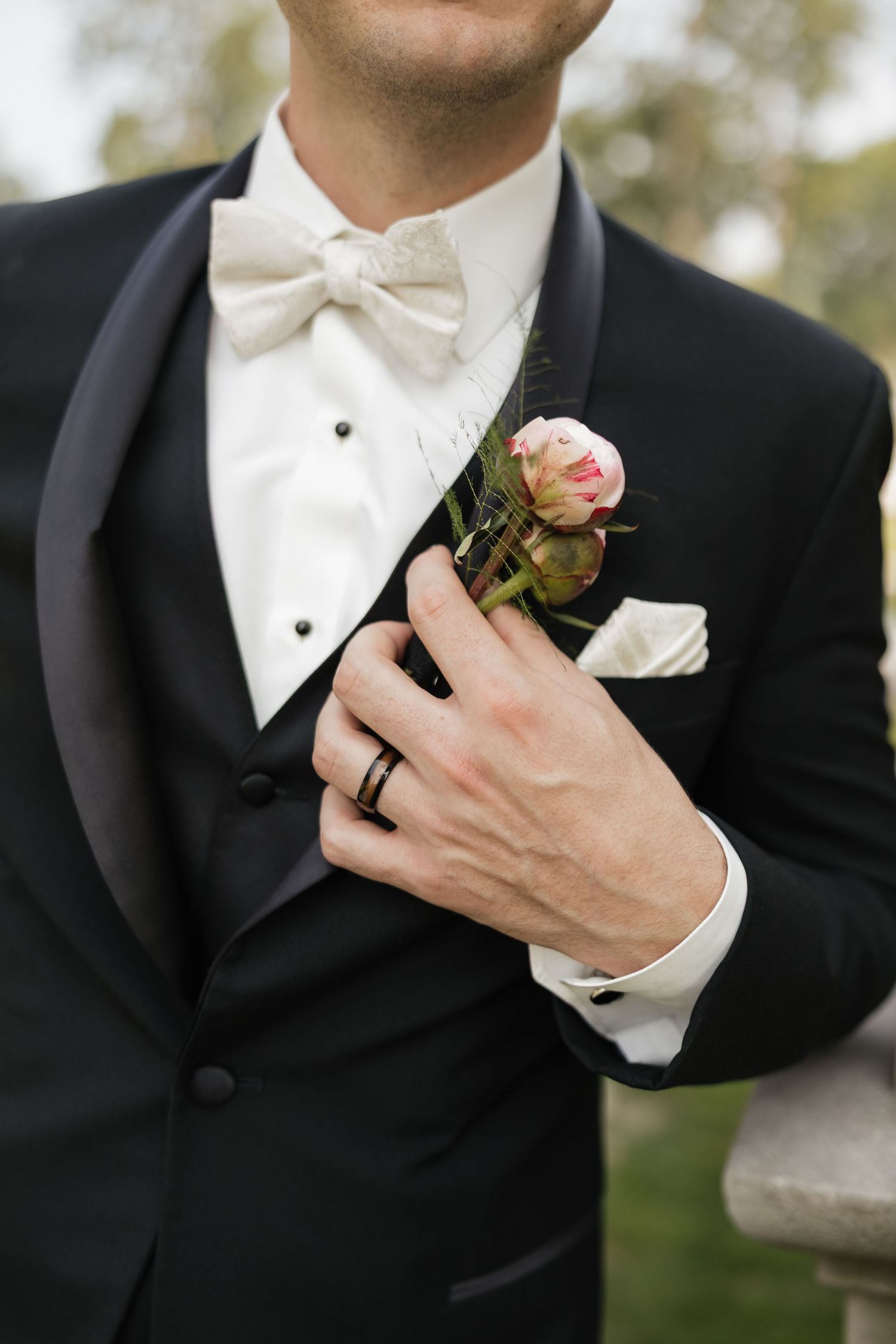 Close-up of groom in black tuxedo with white bow tie adjusting pink rose boutonniere, wearing wedding band.