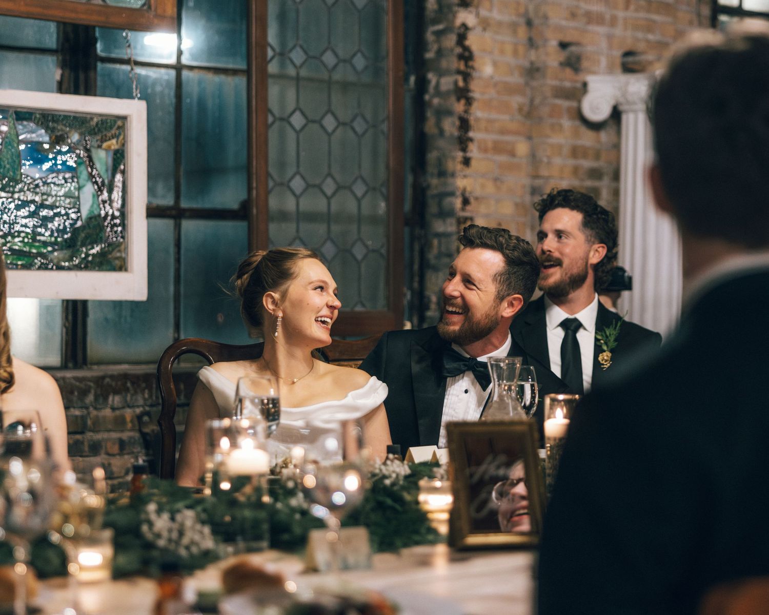 Guests mingle and laugh during an evening celebration at a rustic brick-walled venue with warm lighting.