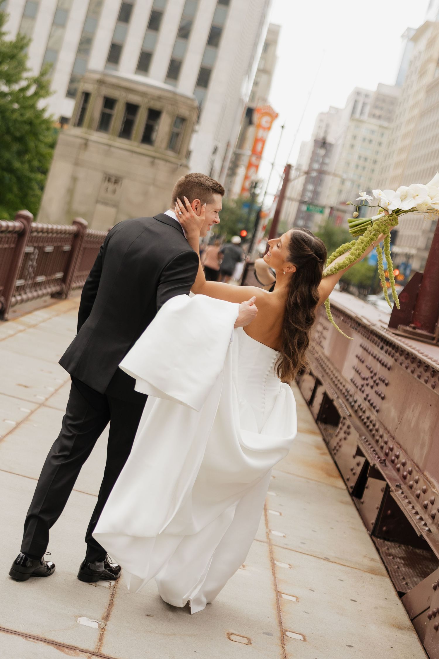 A bride and groom share a romantic dip kiss on a city bridge, she holds a white bouquet in her elegant strapless gown.