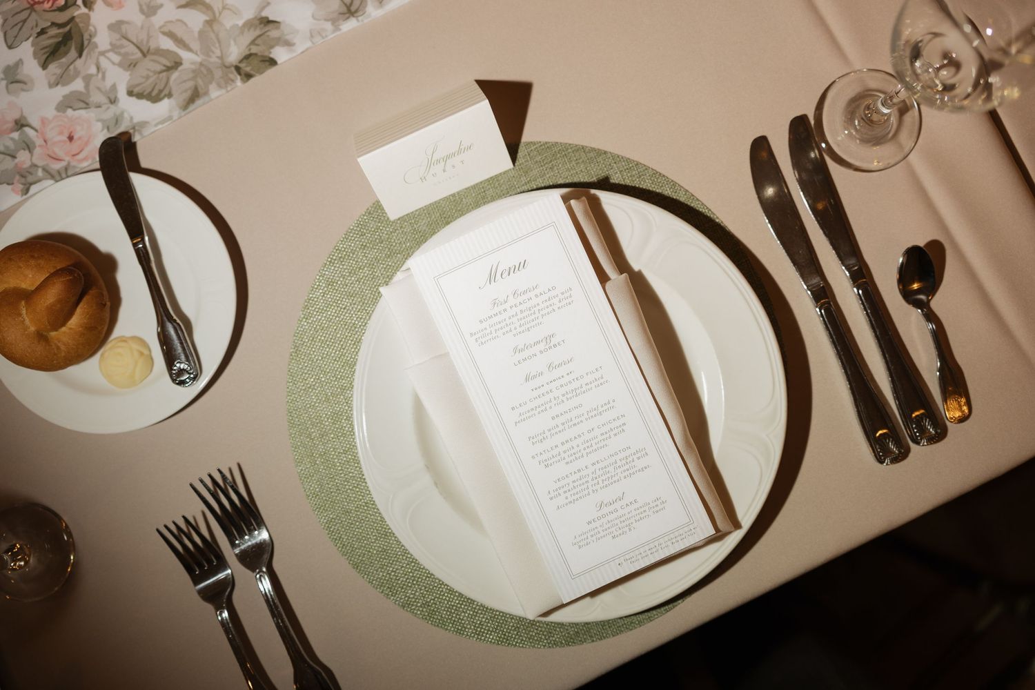 Elegant wedding place setting with menu card, green charger plate, bread roll, and polished silverware on a neutral tablecloth.