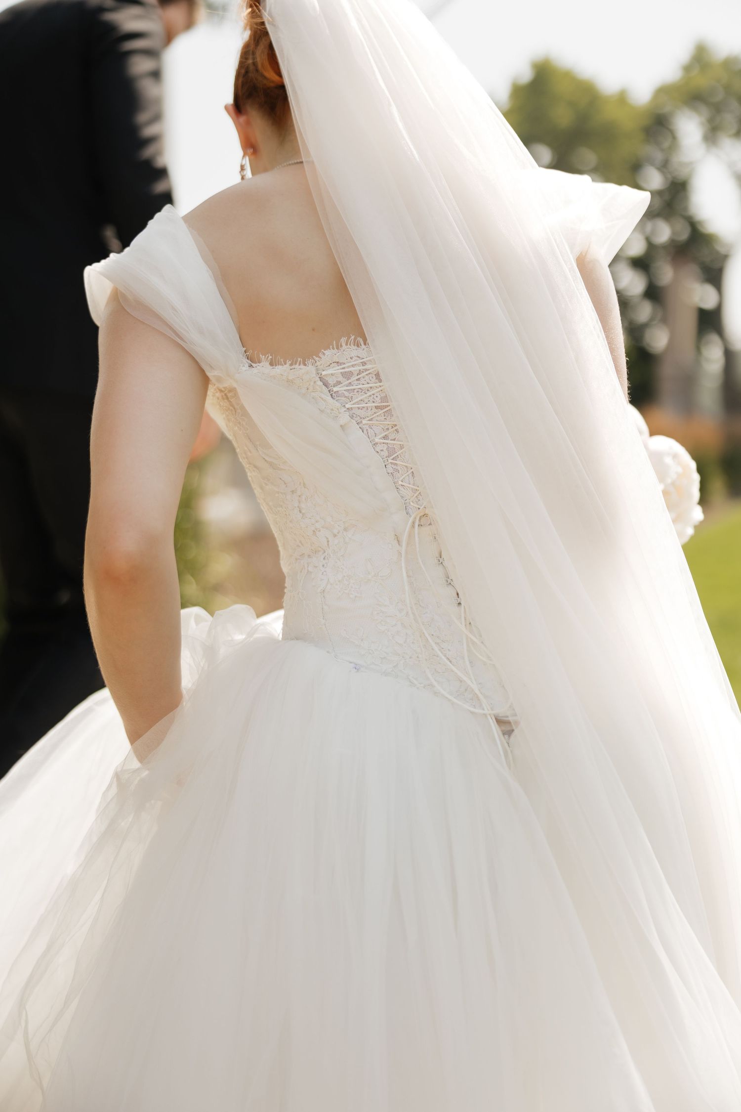 Back view of bride in white lace corset wedding gown with flowing veil and tulle skirt outdoors.