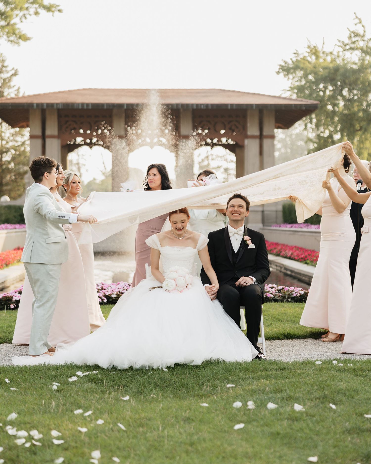 Bride and groom sit on grass while wedding party tosses floral veil at elegant outdoor garden venue.