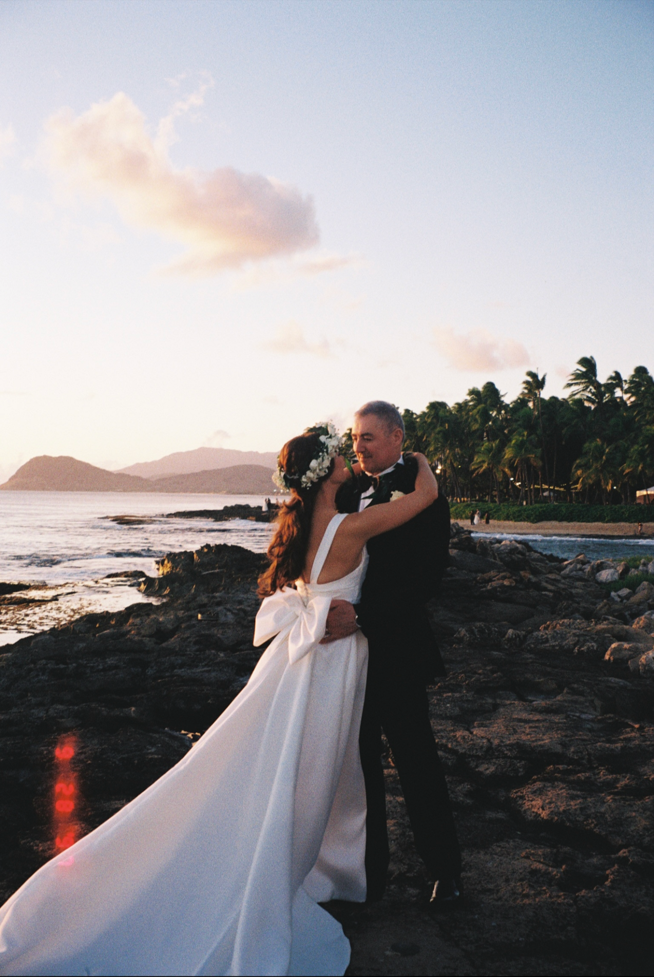 A romantic sunset wedding photoshoot on a rocky Hawaiian beach with palm trees and mountains in the background.