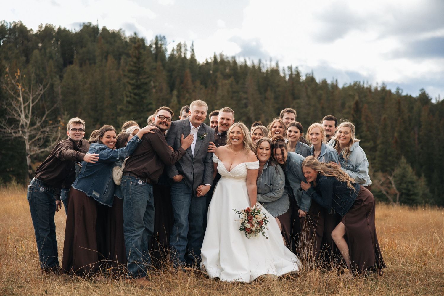 Wedding group photo with guests posing playfully in mountain meadow setting.