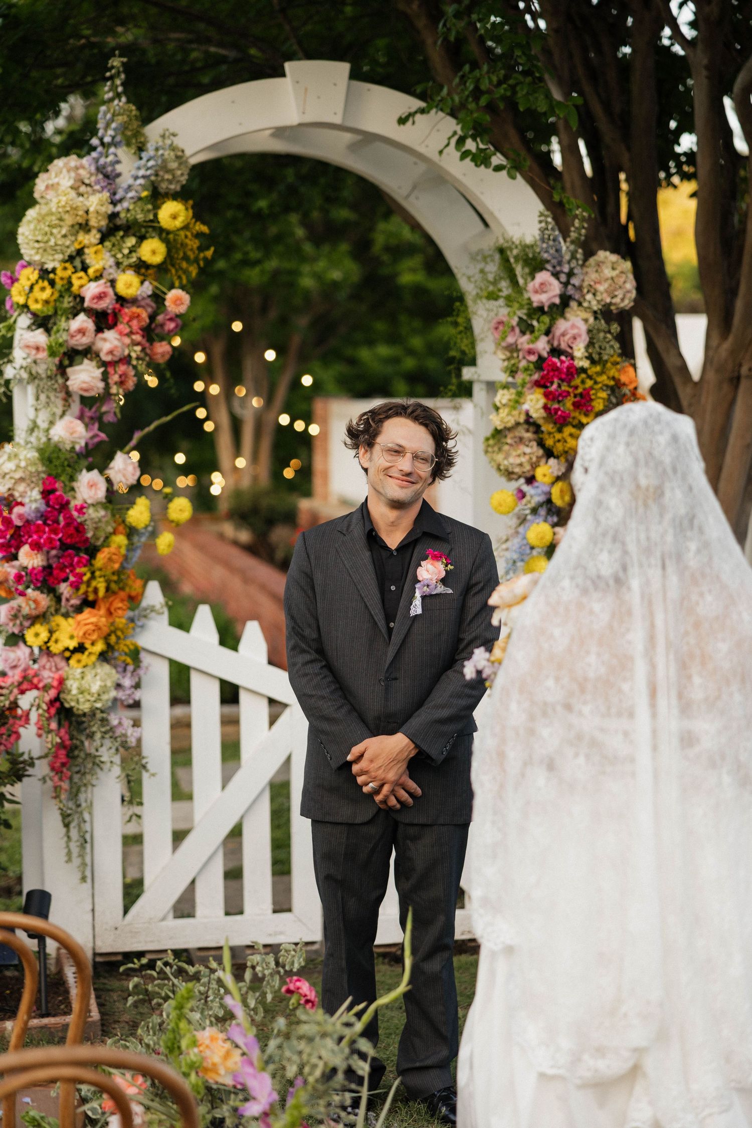 Groom smiling at bride approaching floral arch at outdoor wedding ceremony with colorful blooms.
