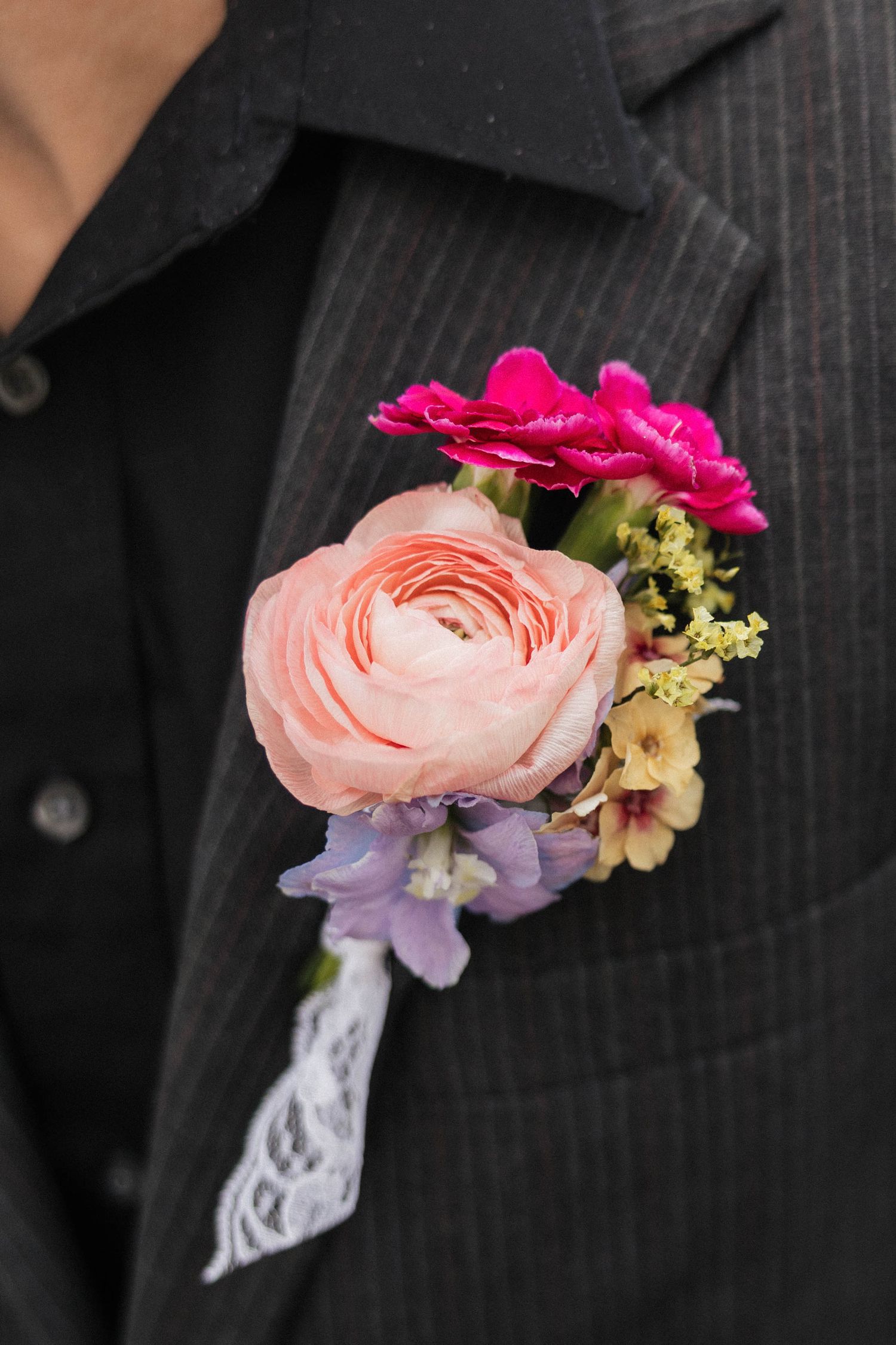 Elegant wedding boutonniere with pink ranunculus, fuchsia roses, and purple flowers on a black pinstripe suit lapel.