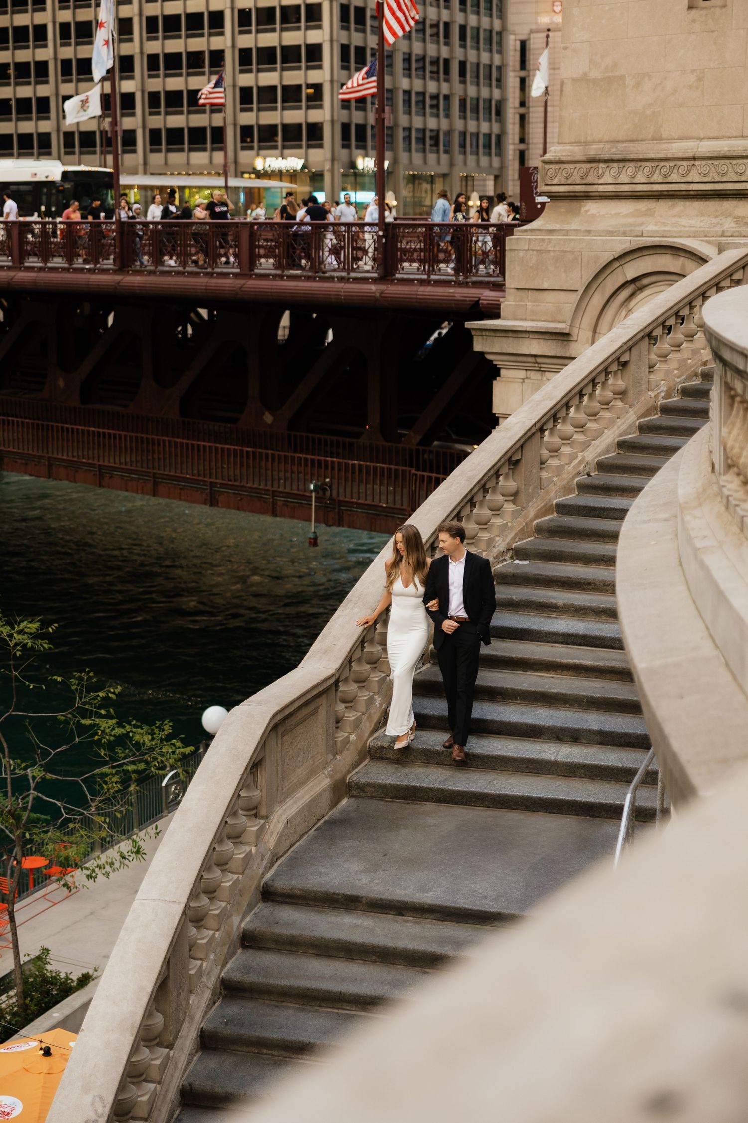 Couple in wedding attire walking down stone stairs along Chicago Riverwalk with bridge and city buildings in background.