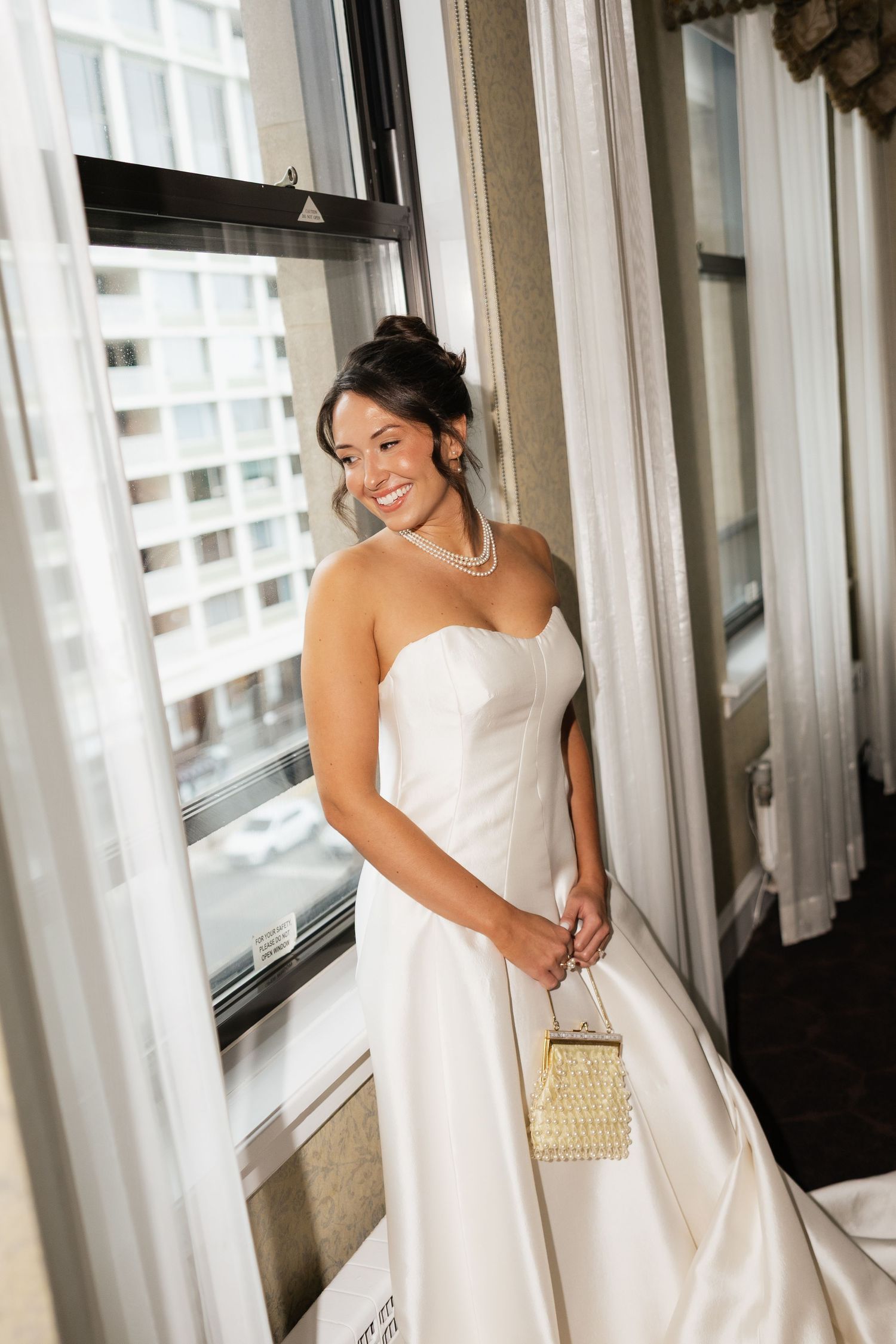Smiling bride in strapless white satin gown holding gold clutch, standing by window with city view.