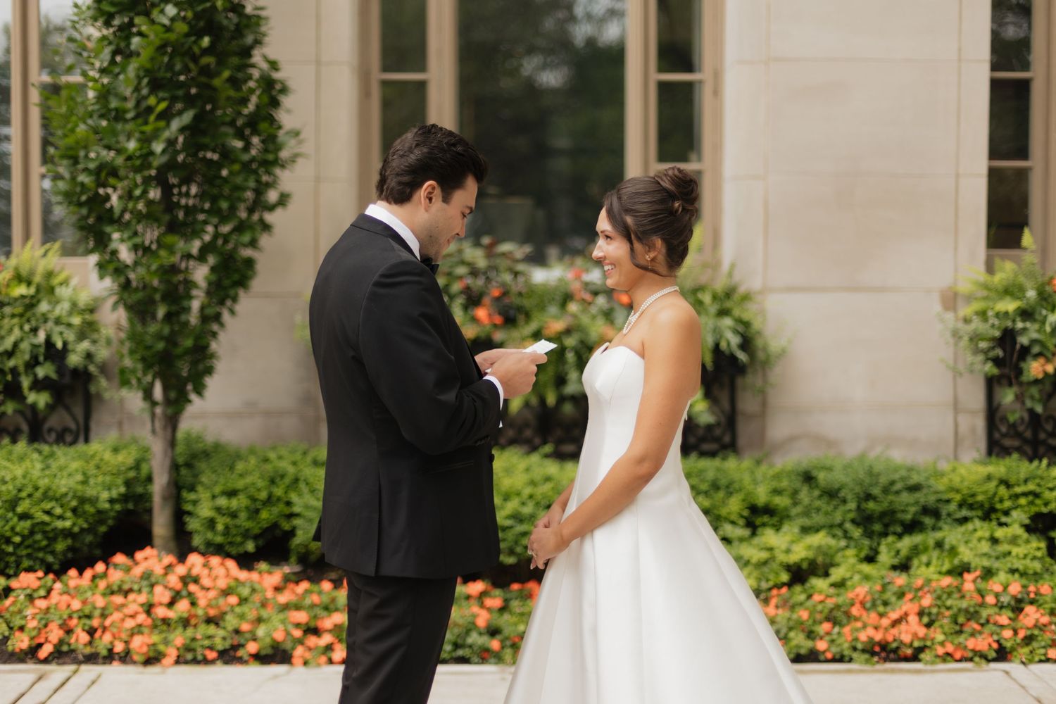 Groom reads vows to smiling bride in white gown outdoors, with lush greenery and orange flowers in background.