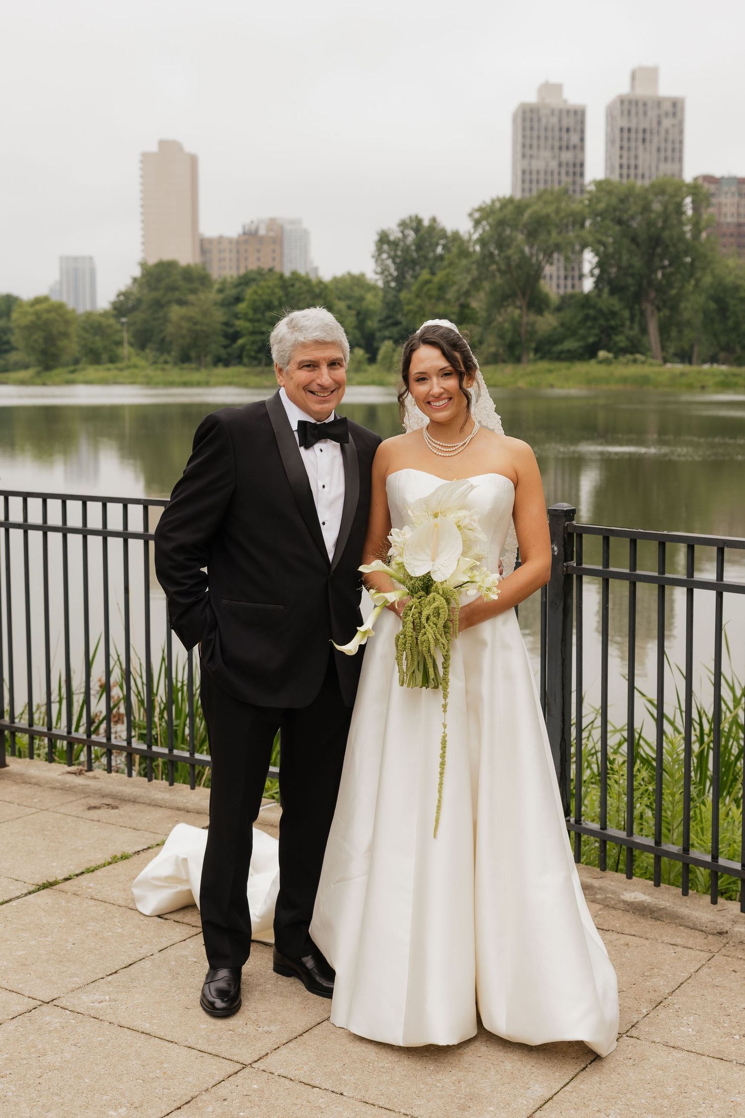 Bride in white gown holding calla lily bouquet poses with man in tuxedo by Central Park lake with city skyline backdrop.