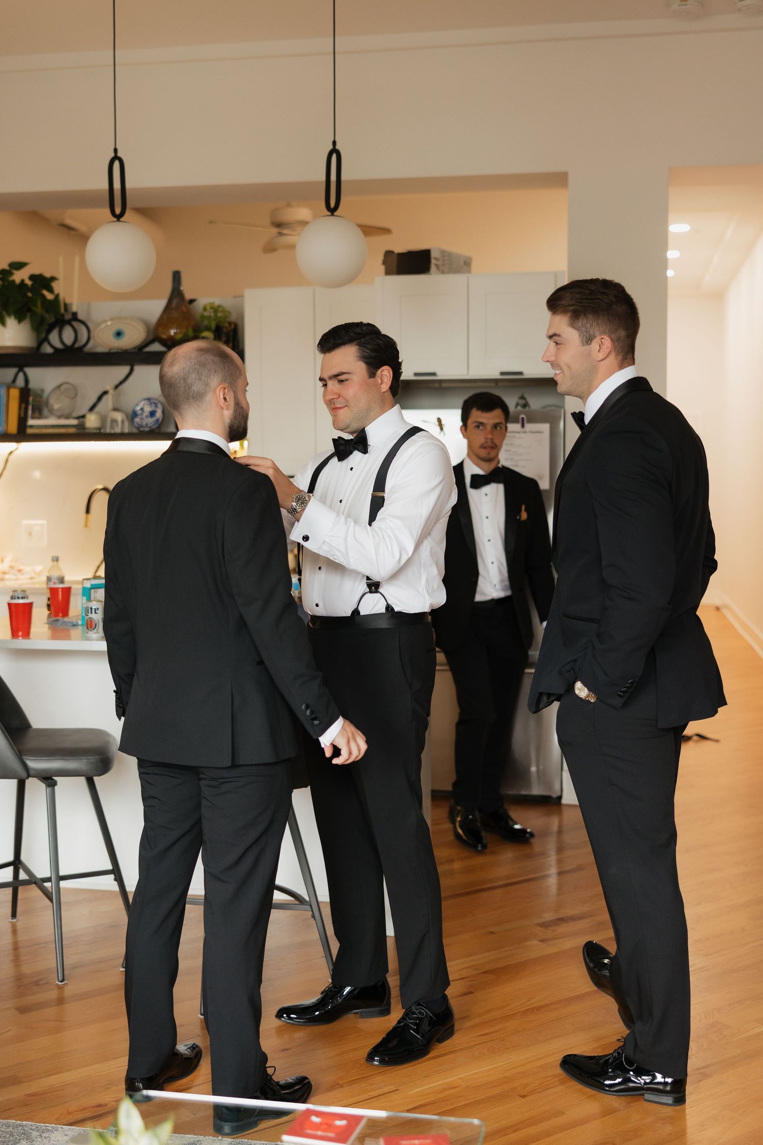 Groomsmen in black tuxedos with bow ties help each other prepare in a modern kitchen before a wedding.