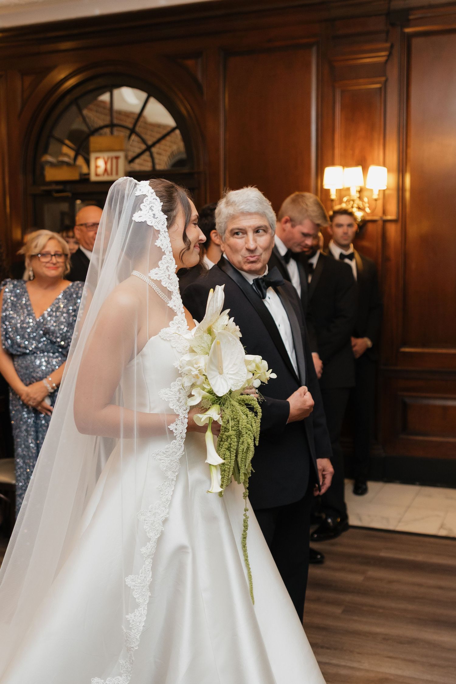 Bride in white gown and veil holds floral bouquet while walking with older man in tuxedo at elegant wedding ceremony.