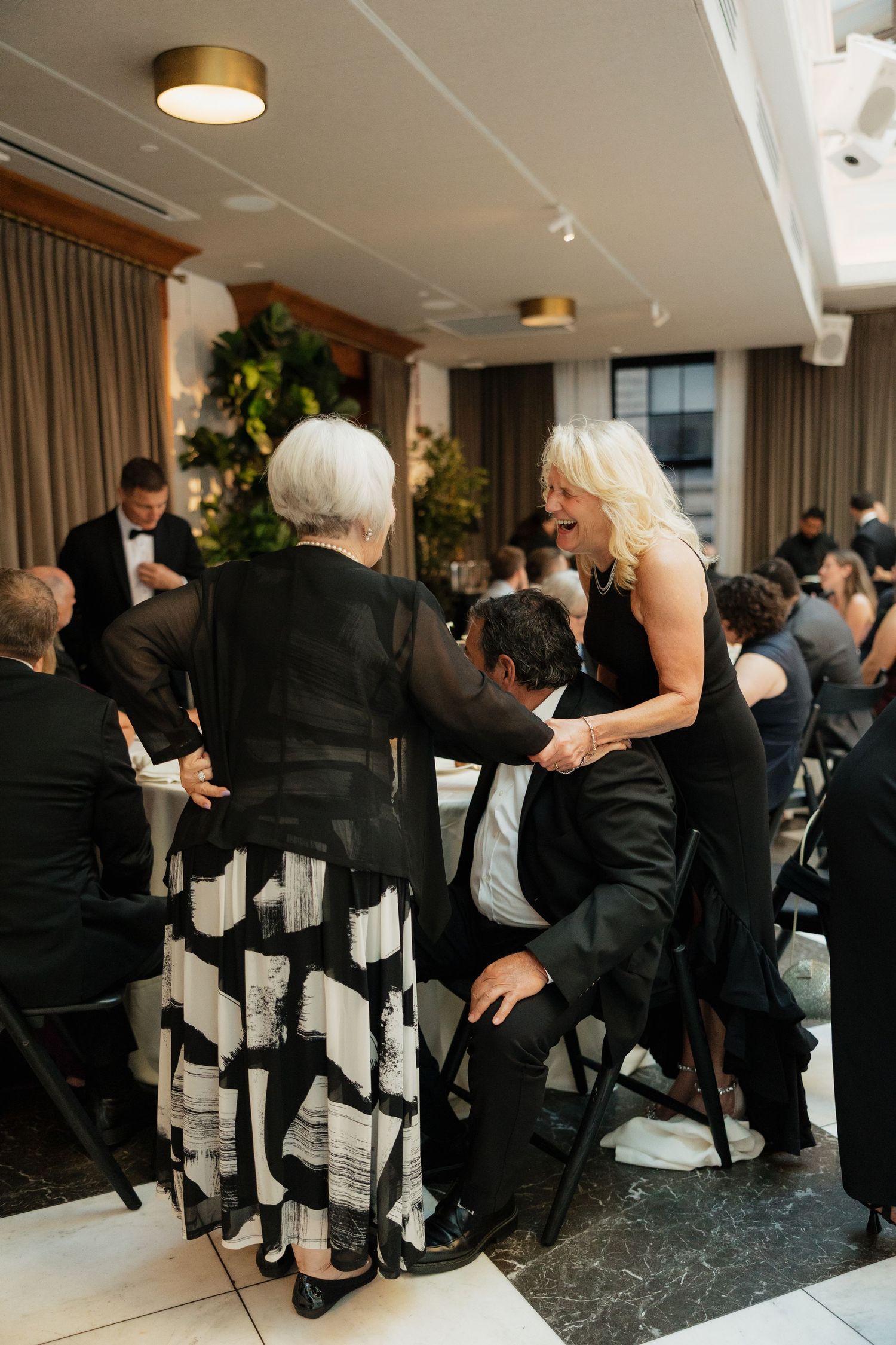 Wedding guests in formal black attire mingle and laugh together during a lively indoor wedding reception dinner.