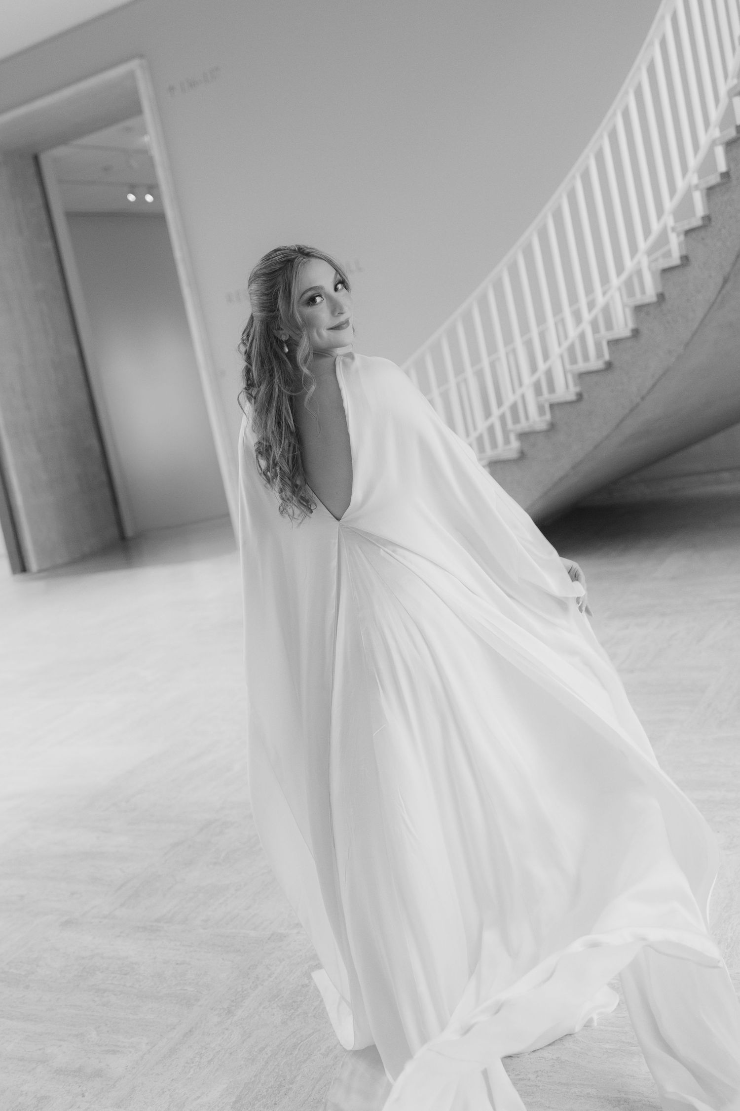 A bride in a flowing white wedding dress twirls elegantly in front of a white staircase in a black and white photo.