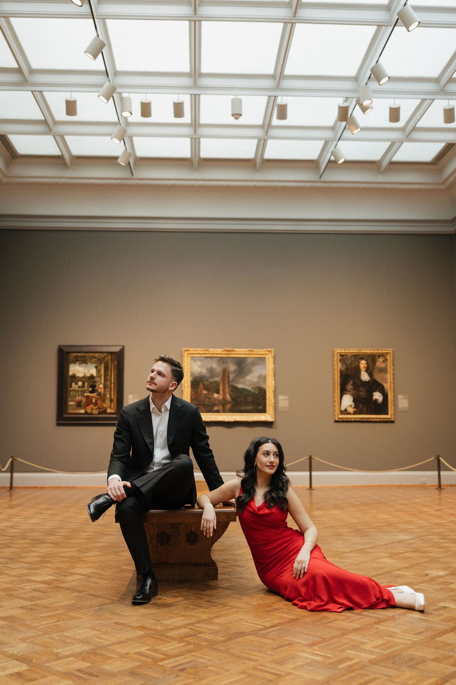 A couple poses playfully in an art gallery with wood floors and paintings on display.