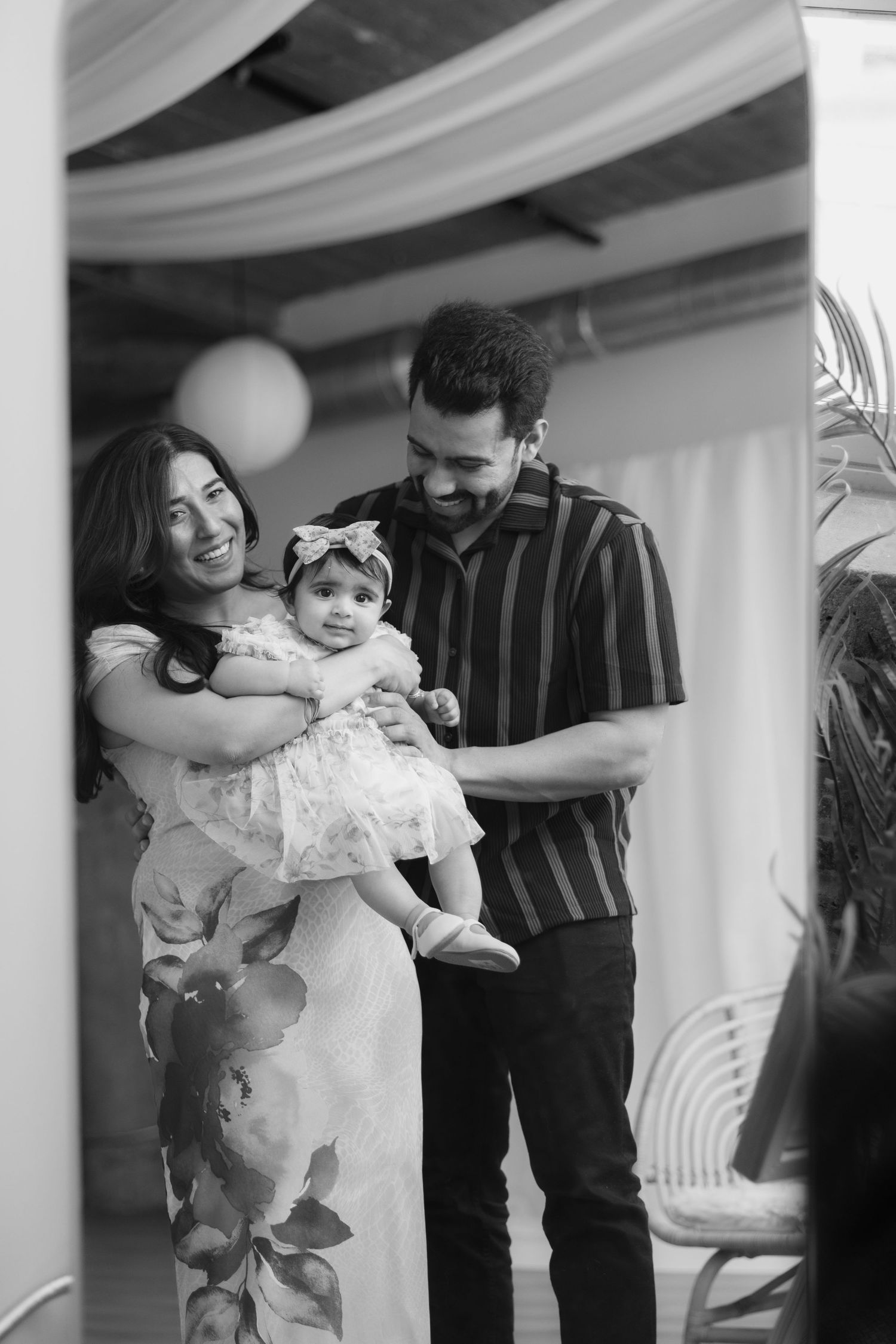 Black and white photo of smiling parents holding their baby girl in a floral dress at a celebration.